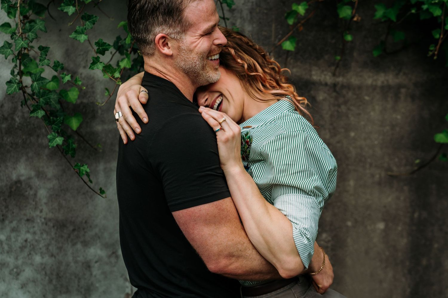A romantic couple embraces and laughs together against a concrete wall with green ivy in the background.