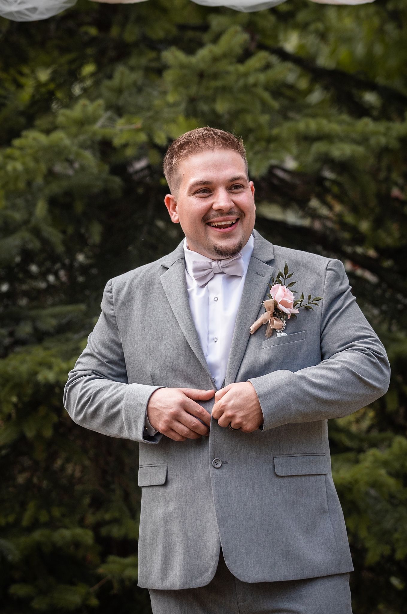 Groom in light grey suit with boutonniere smiling outdoors.