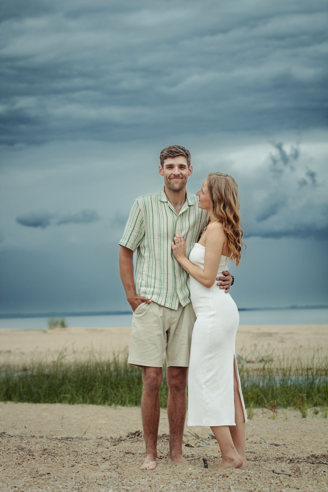 A couple shares an intimate moment on a beach with dramatic stormy clouds overhead.