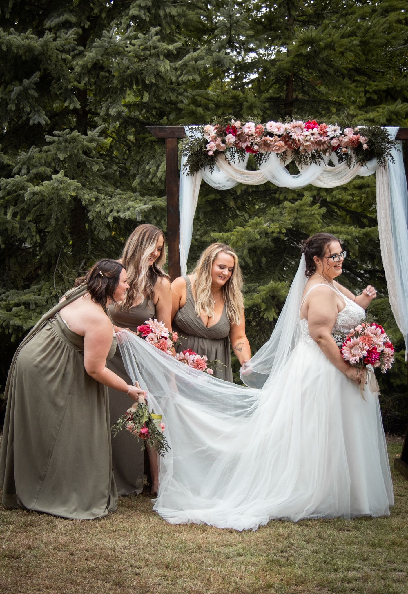 Bridesmaids in olive dresses help adjust the train of a wedding dress under a floral arch.