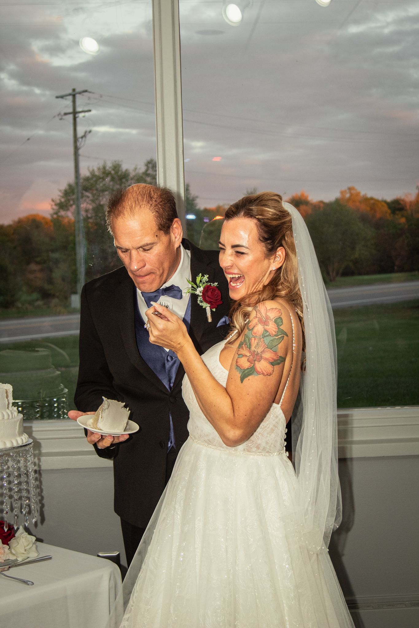 A couple shares a joyful moment during their wedding reception in a venue with large windows at sunset.