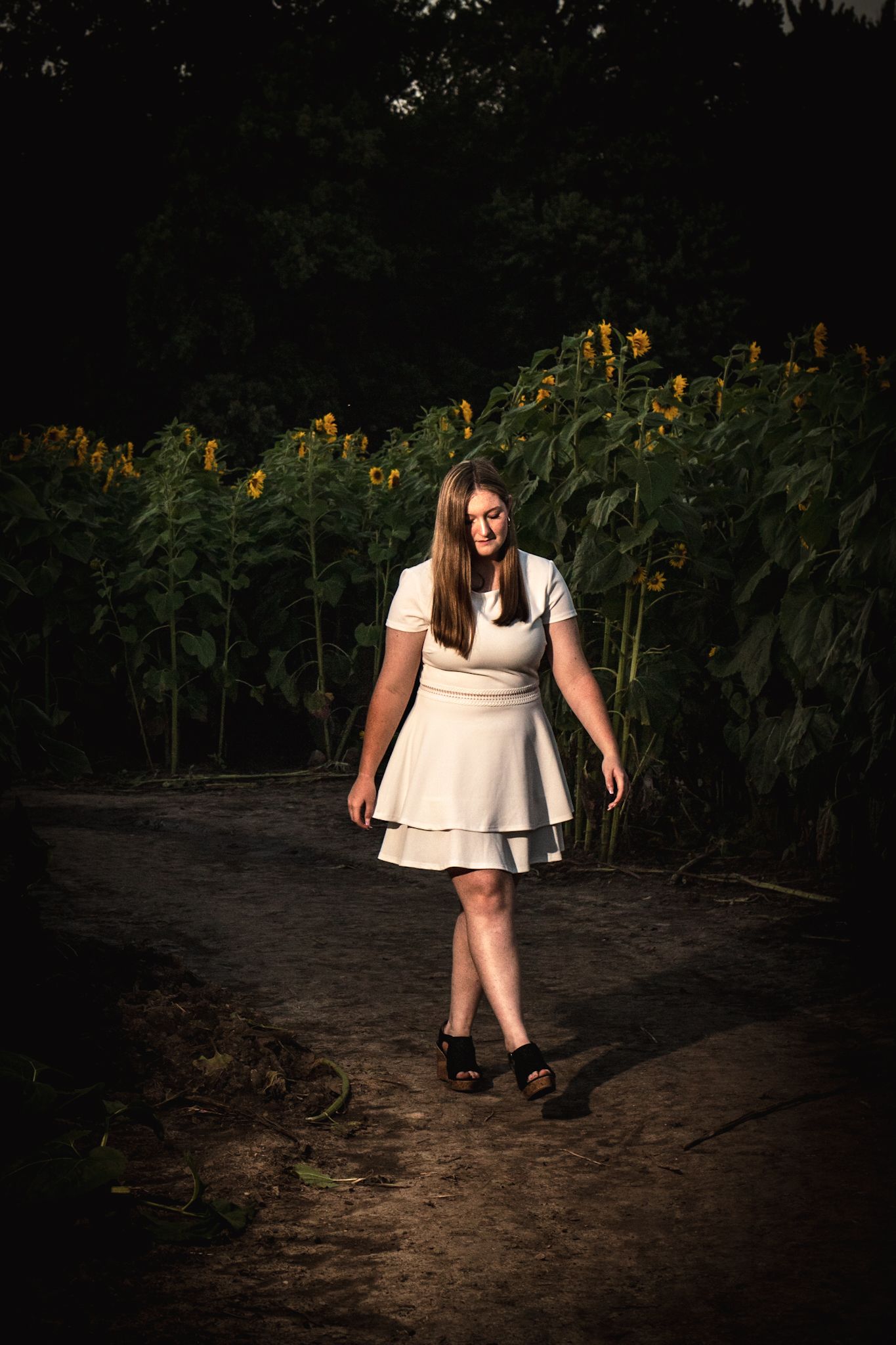 A person in a white dress walks along a dark path with sunflowers in the background.