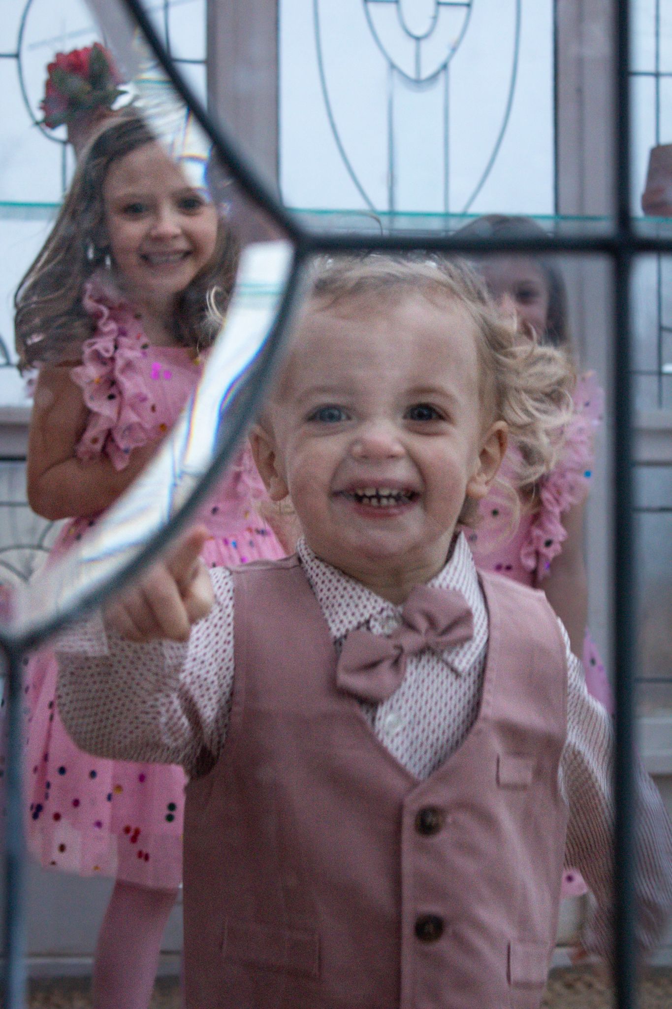 Young children in festive pink attire share joyful smiles in front of a decorative glass window.
