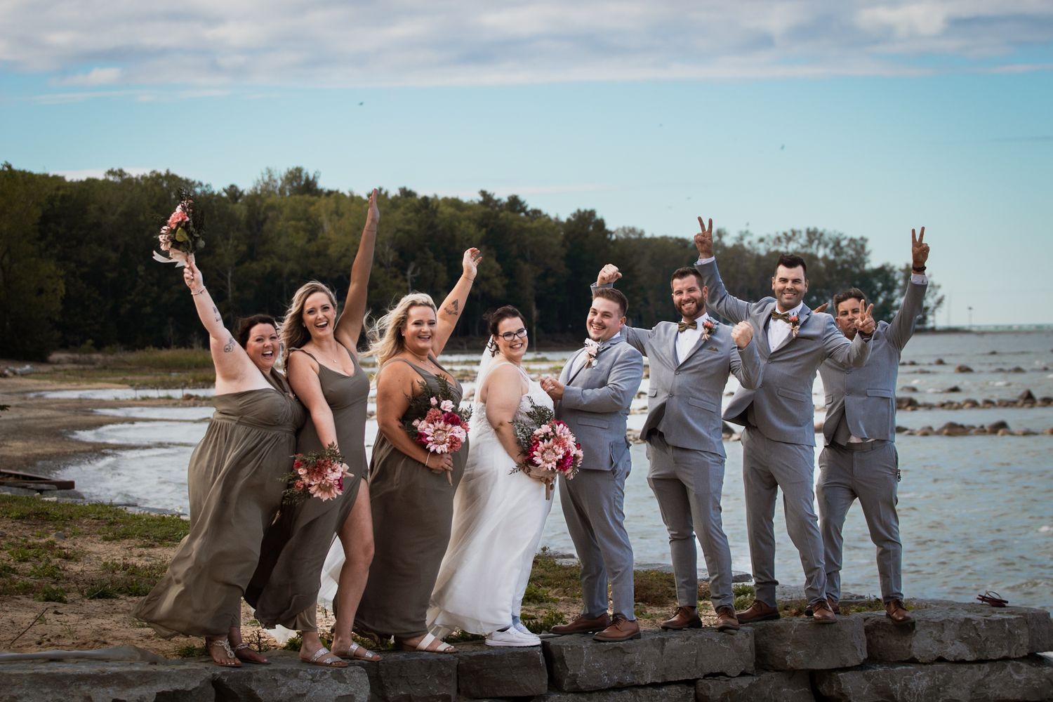 Wedding party celebrates joyfully on a scenic beach with arms raised against blue sky and ocean backdrop.