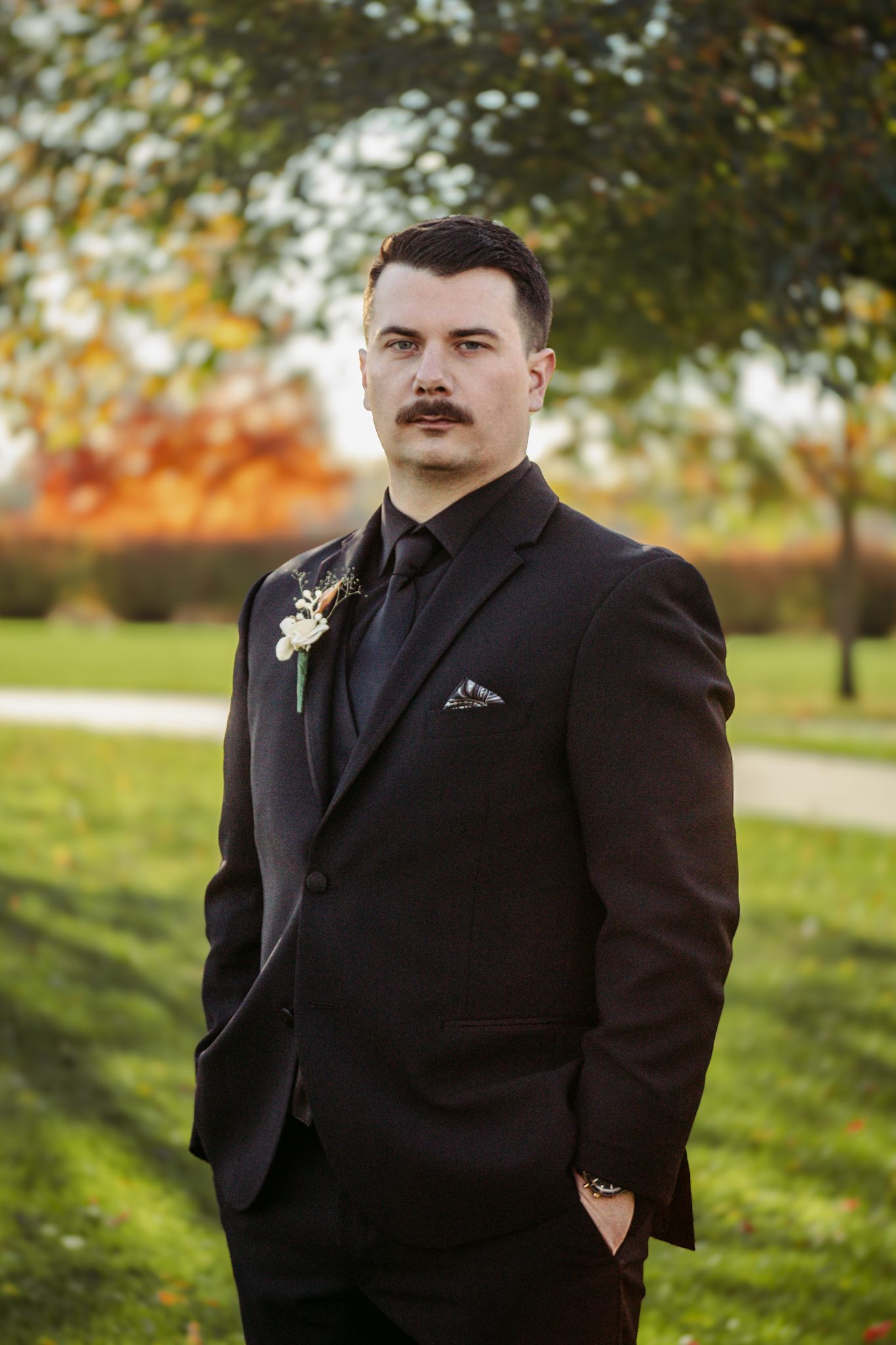 A formal portrait of a groom in a black suit with boutonniere against an autumn background.