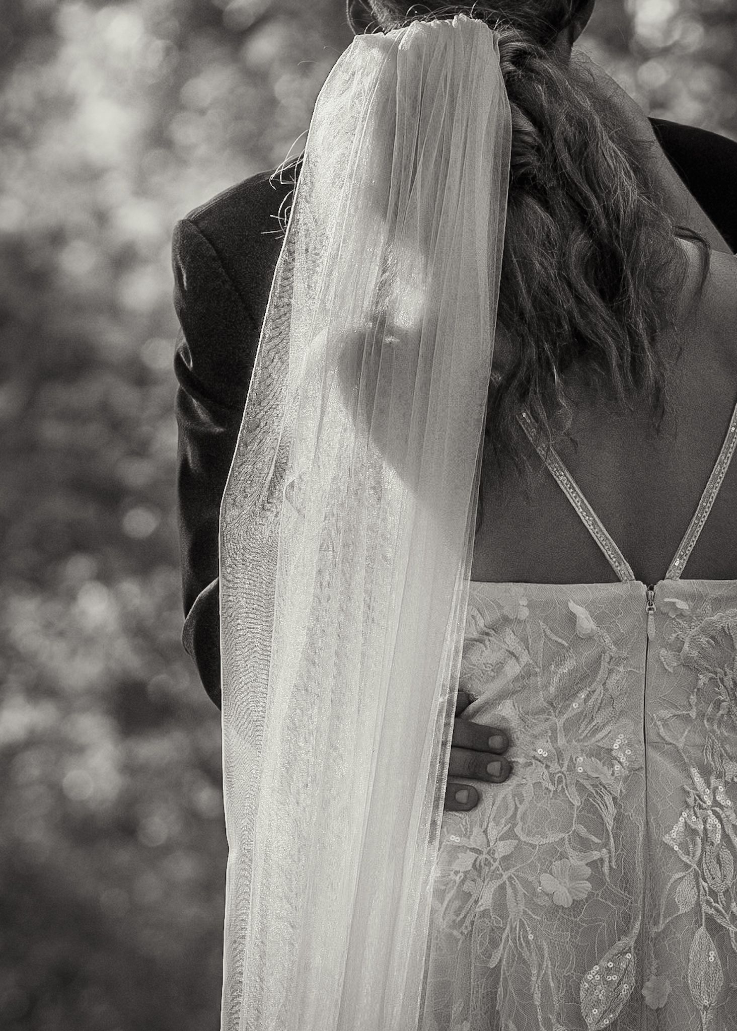A delicate bridal veil flows down in black and white against an embroidered wedding dress with a bokeh background.