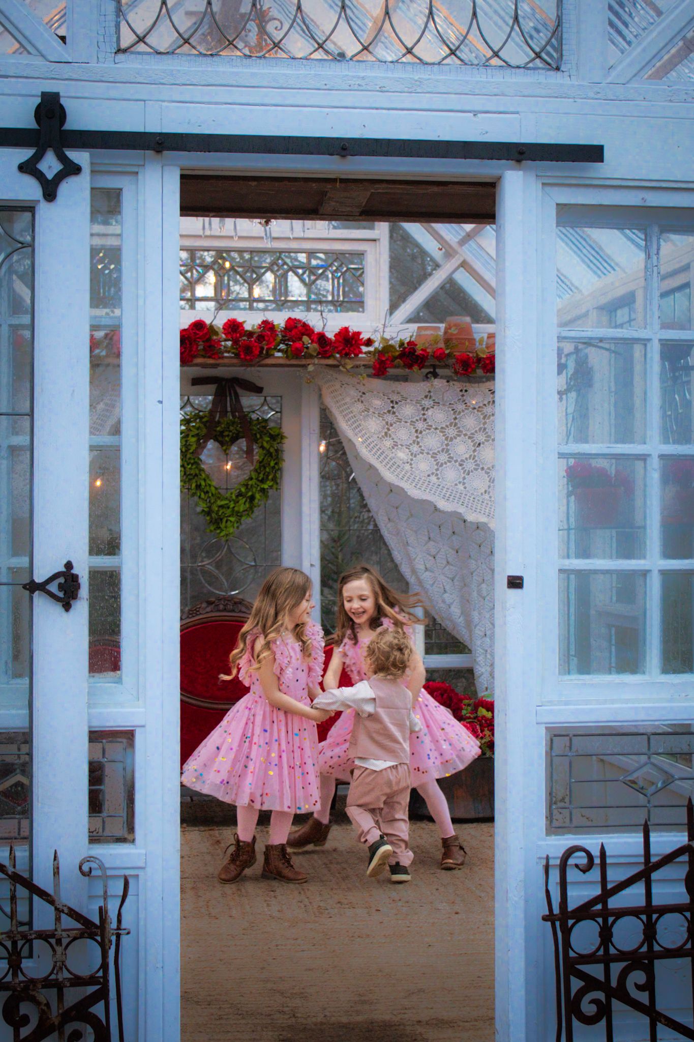Children in pink dresses dancing through doorway decorated with holiday wreath and garland.