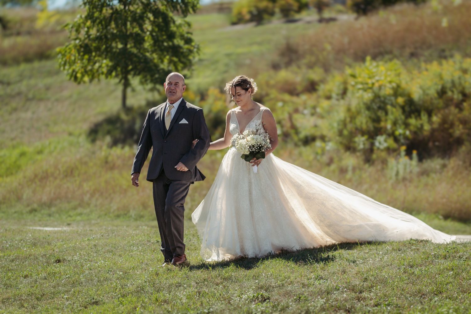 A newlywed couple walks through a grassy hillside on their wedding day.