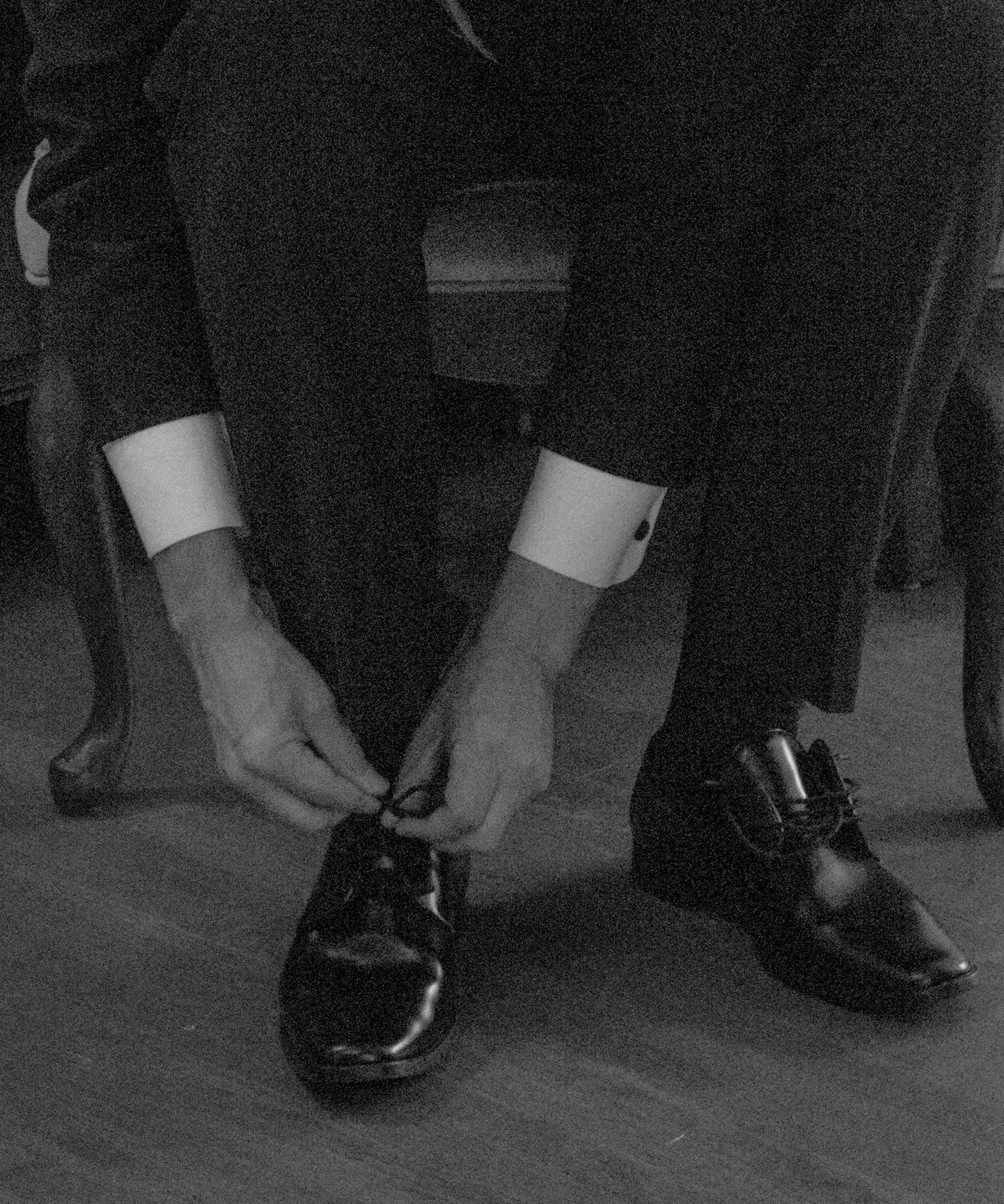 Person tying black formal dress shoes on wooden floor.