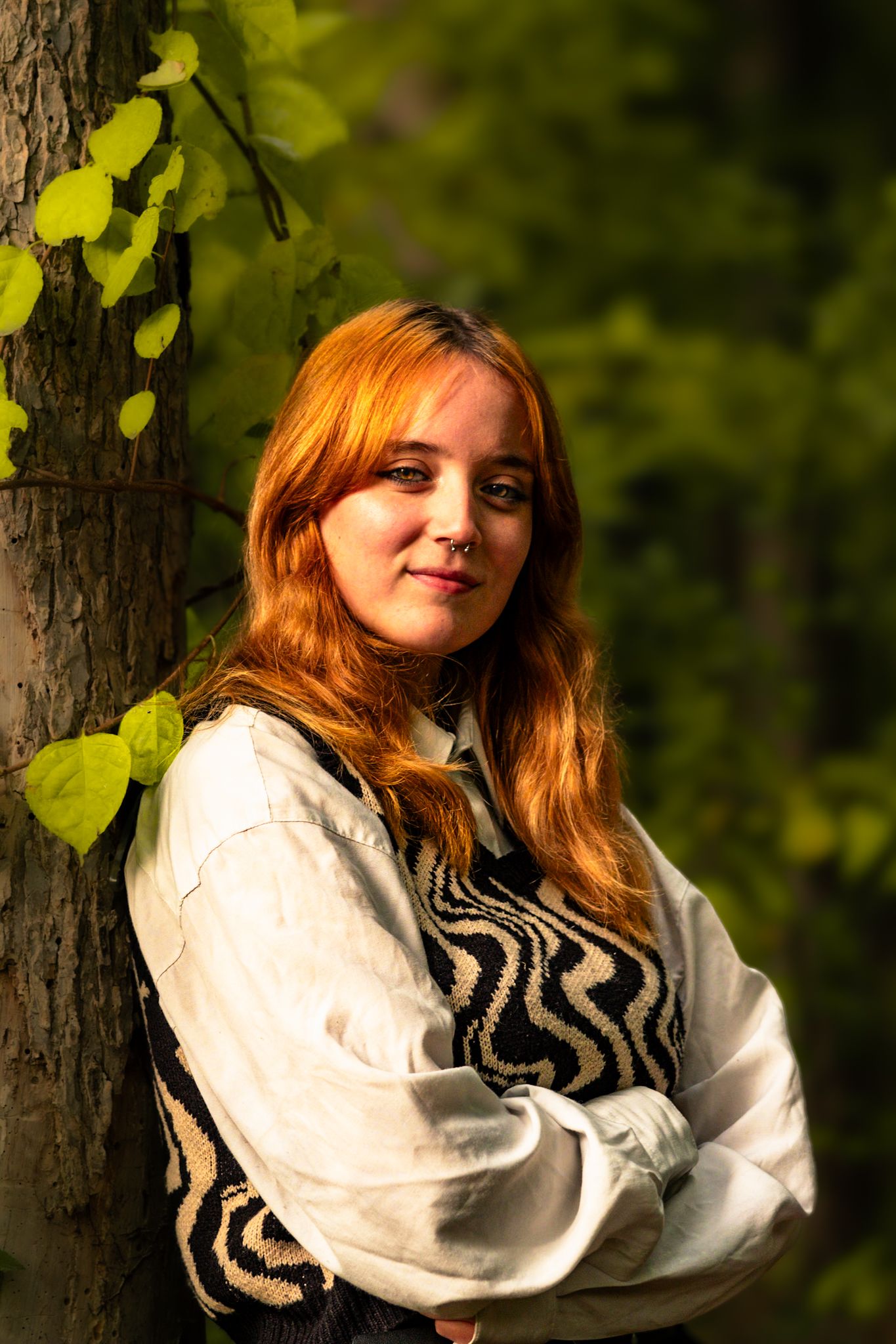 A person in zigzag pattern sweater leans against tree trunk with sunlit leaves in background.