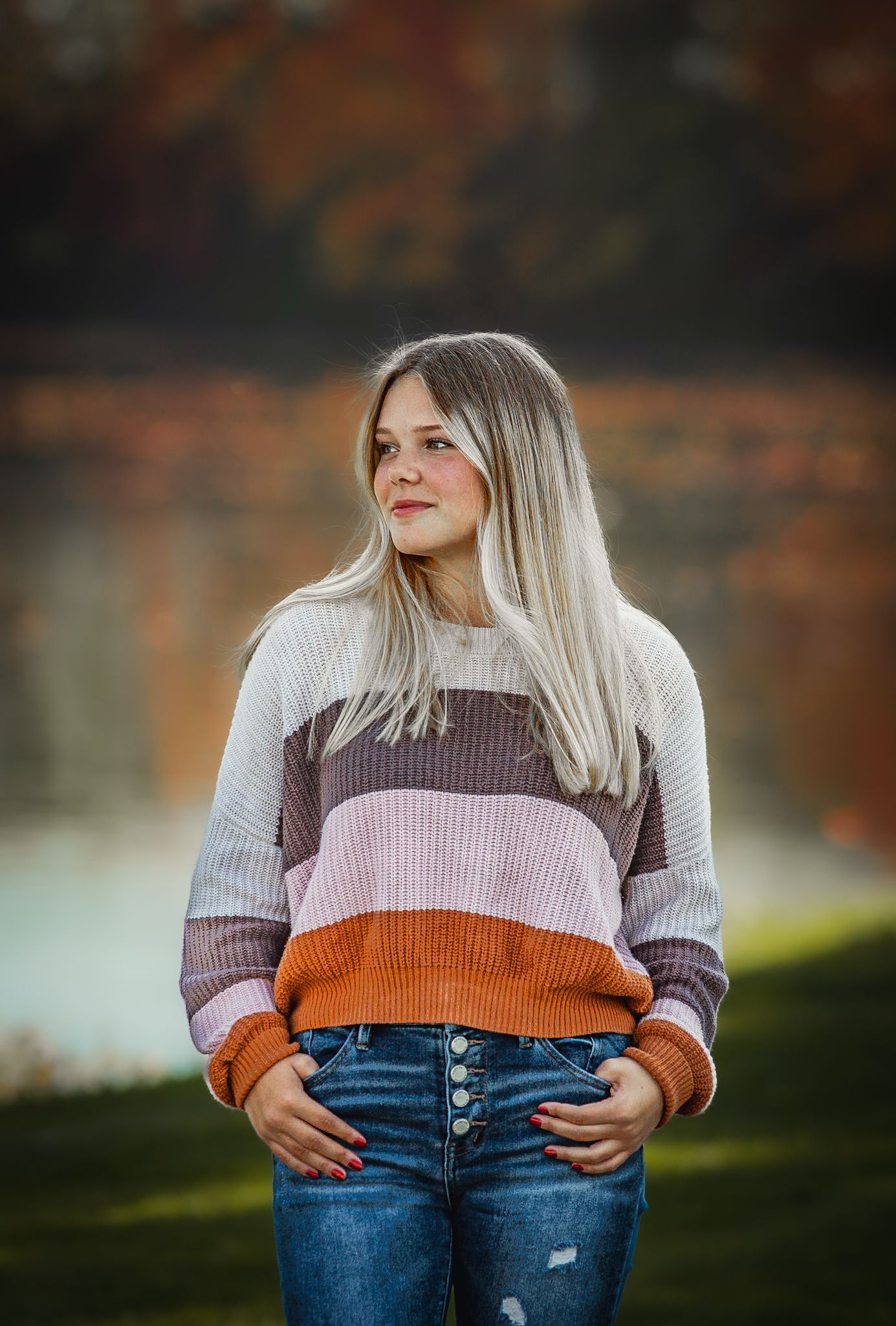 A person in a colorful striped sweater and jeans poses outdoors during autumn.