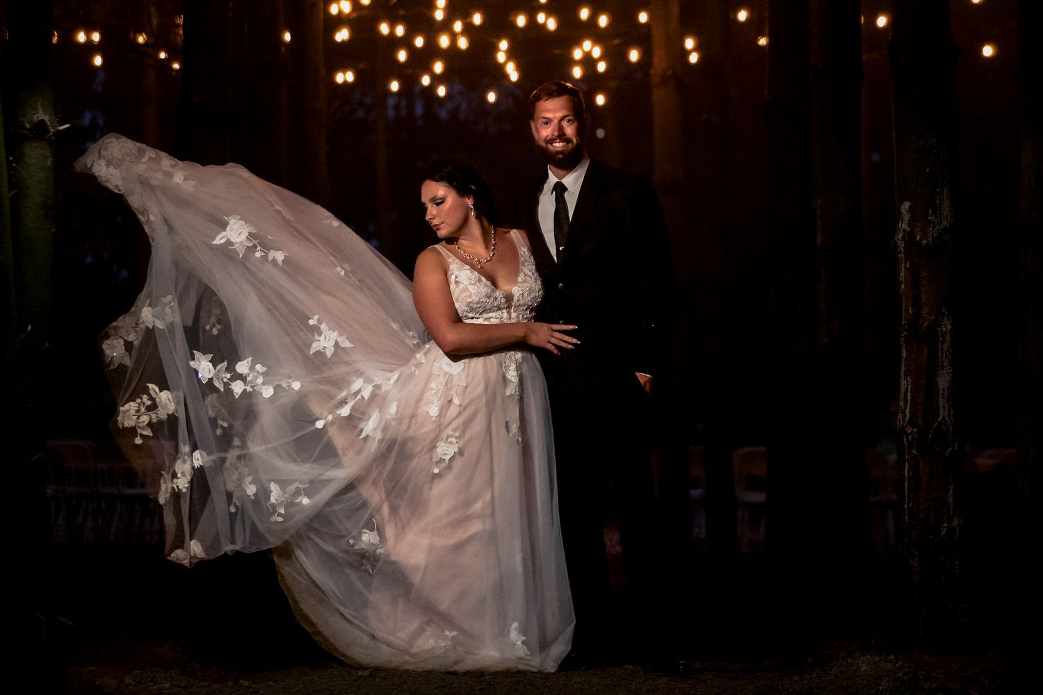 A bride in a flowing white wedding dress is lifted and twirled during a romantic dance moment in a dimly lit venue.
