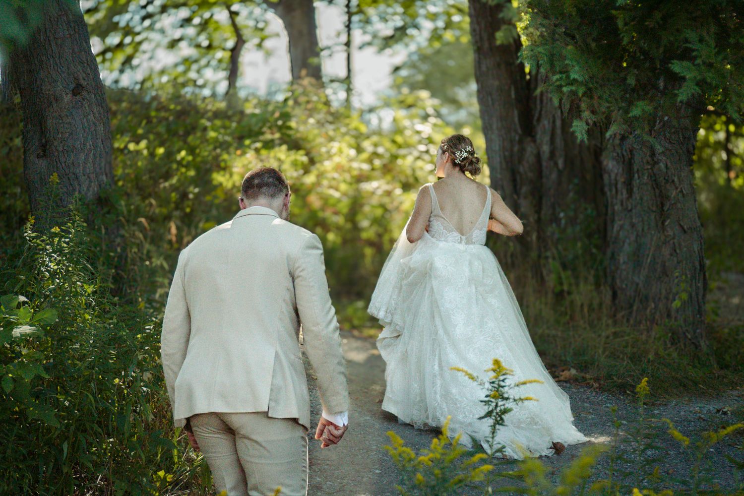 A couple walks down a sunlit path in wedding attire with the bride's dress trailing behind at The Cathedral Barn in Traverse City Michigan.