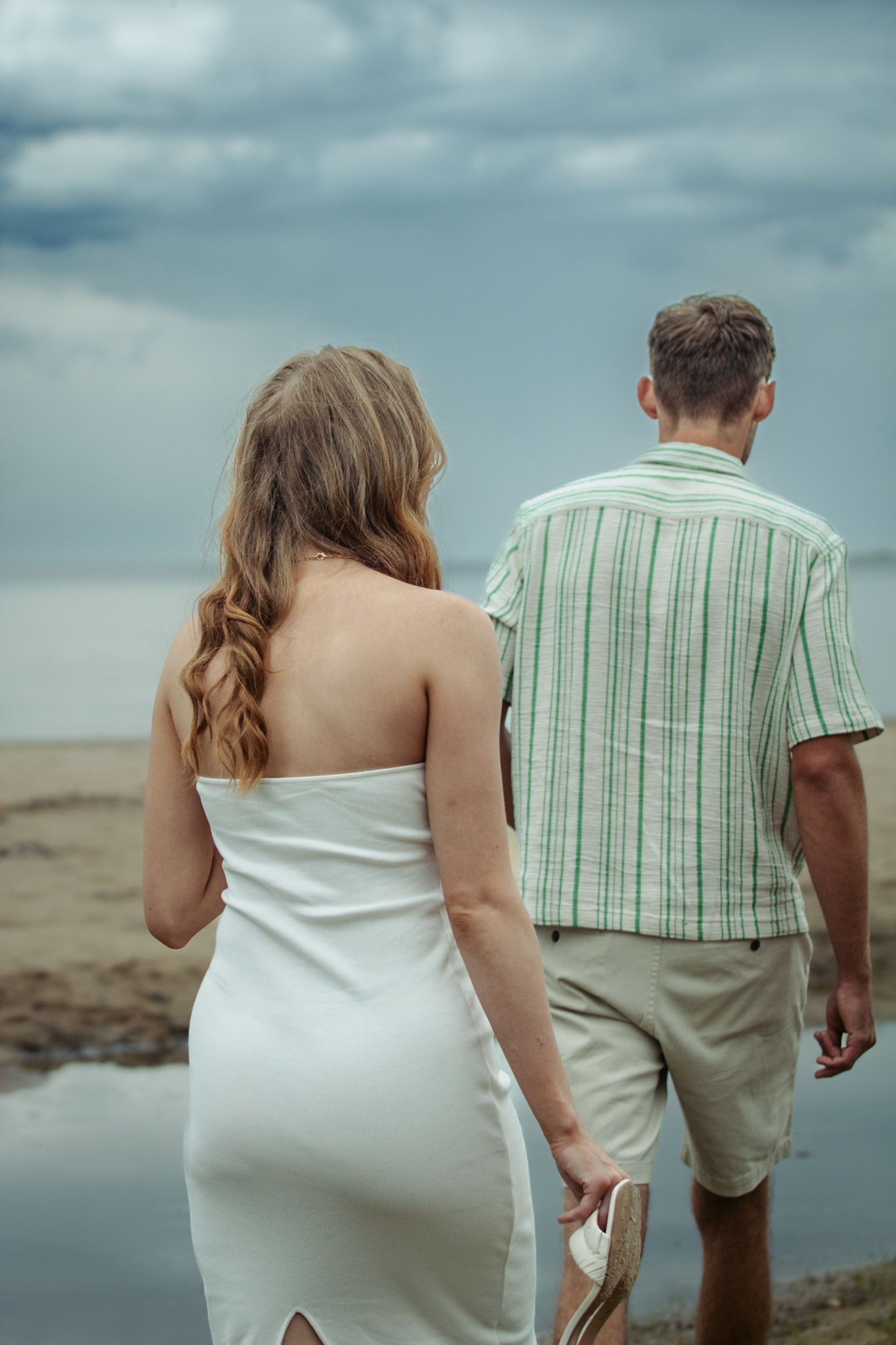 A couple walks along the beach wearing casual summer attire against a cloudy sky.