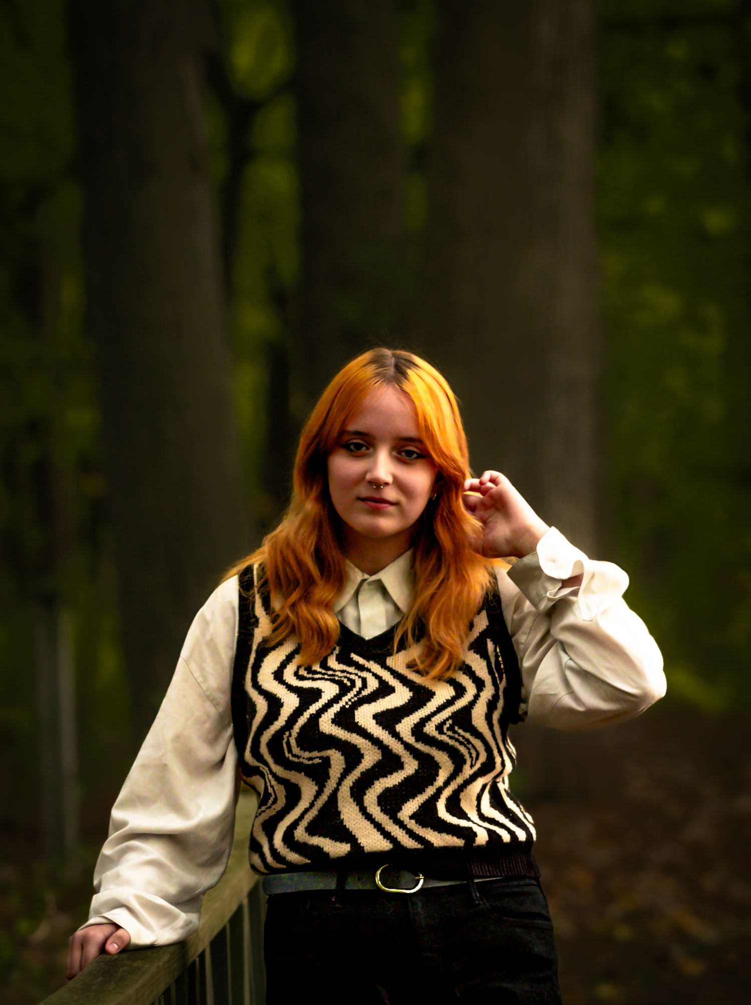A person in a black and white patterned sweater vest poses in a dark forest setting.