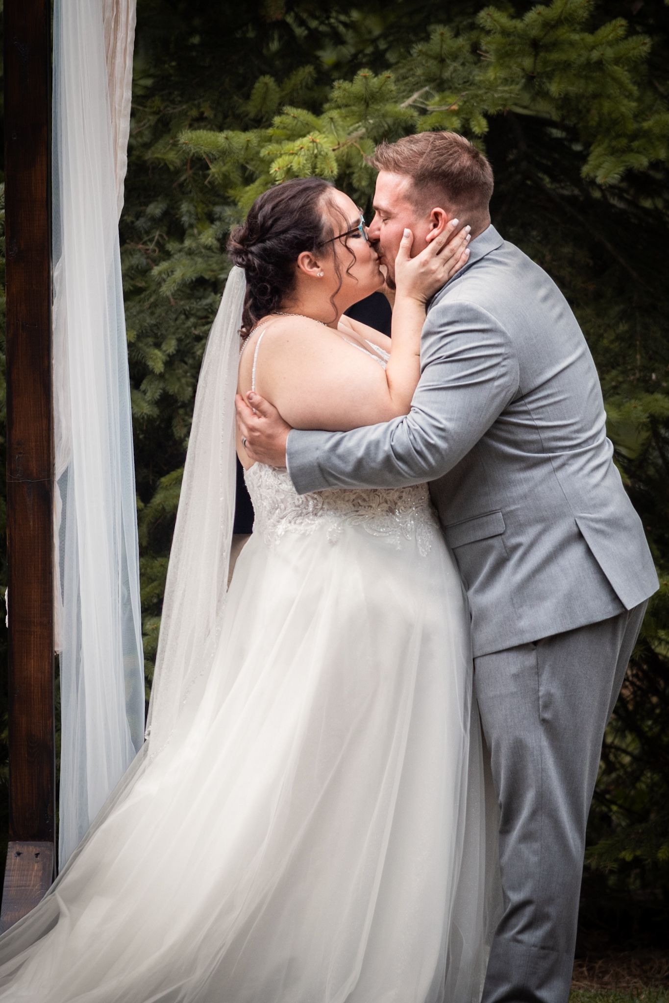 Wedding couple shares first kiss under draped white gazebo surrounded by greenery.