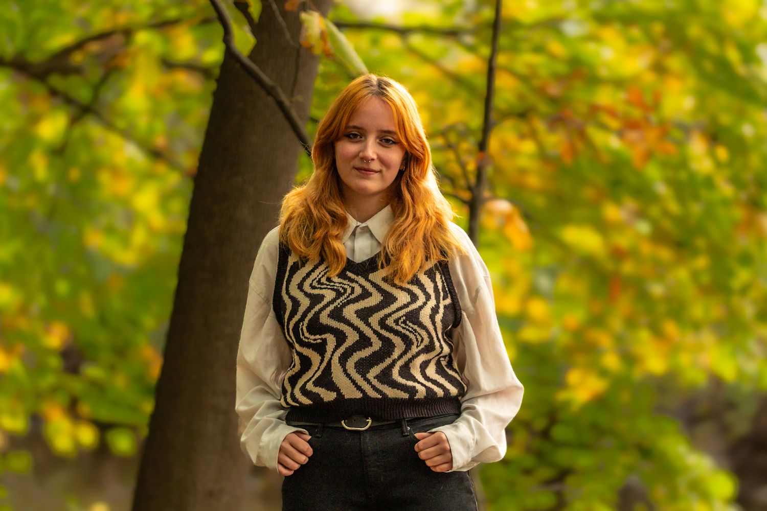 A person wearing a stylish black and white zigzag pattern sweater vest poses outdoors with autumn foliage in background.