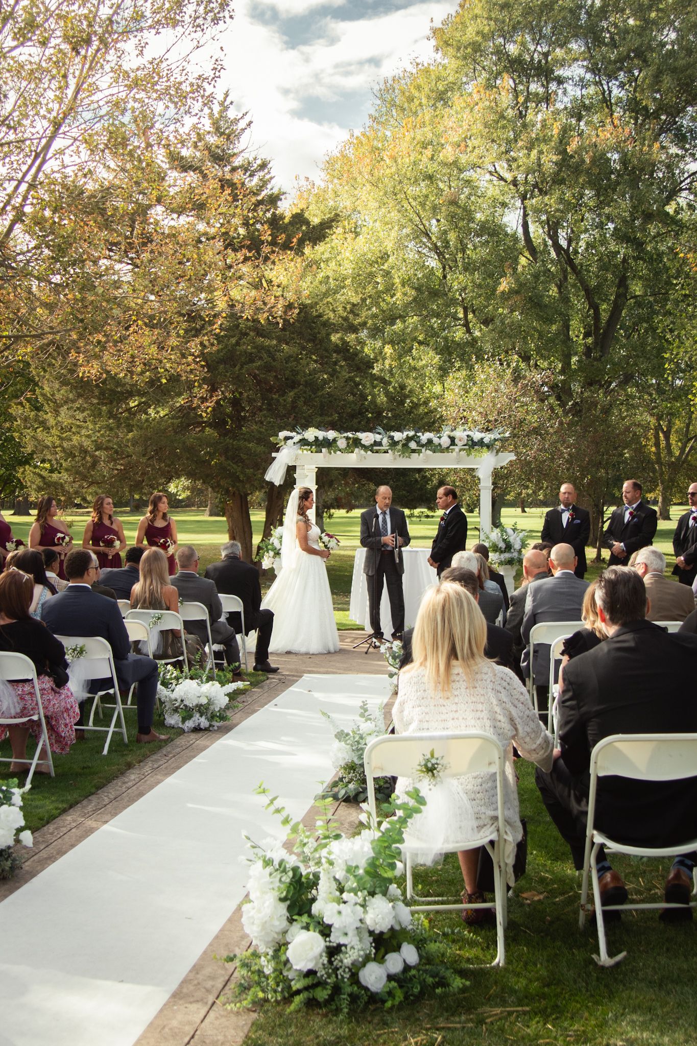 An outdoor wedding ceremony setup with white chairs and floral arrangements along a paved pathway surrounded by autumn trees.
