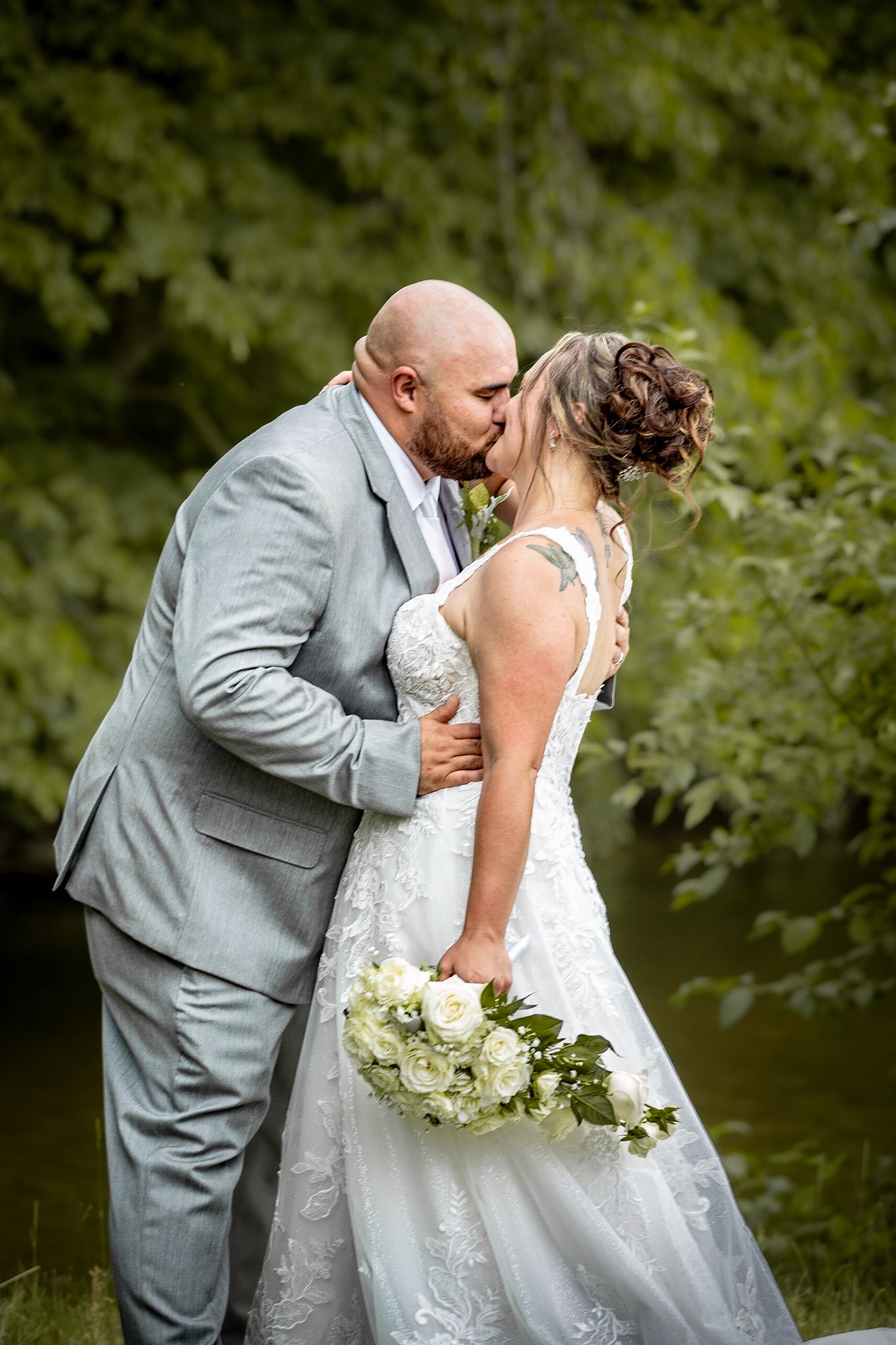 A romantic wedding portrait by a pond with white bouquet and elegant attire at Riverwalk Place venue in Gladwin Michigan.