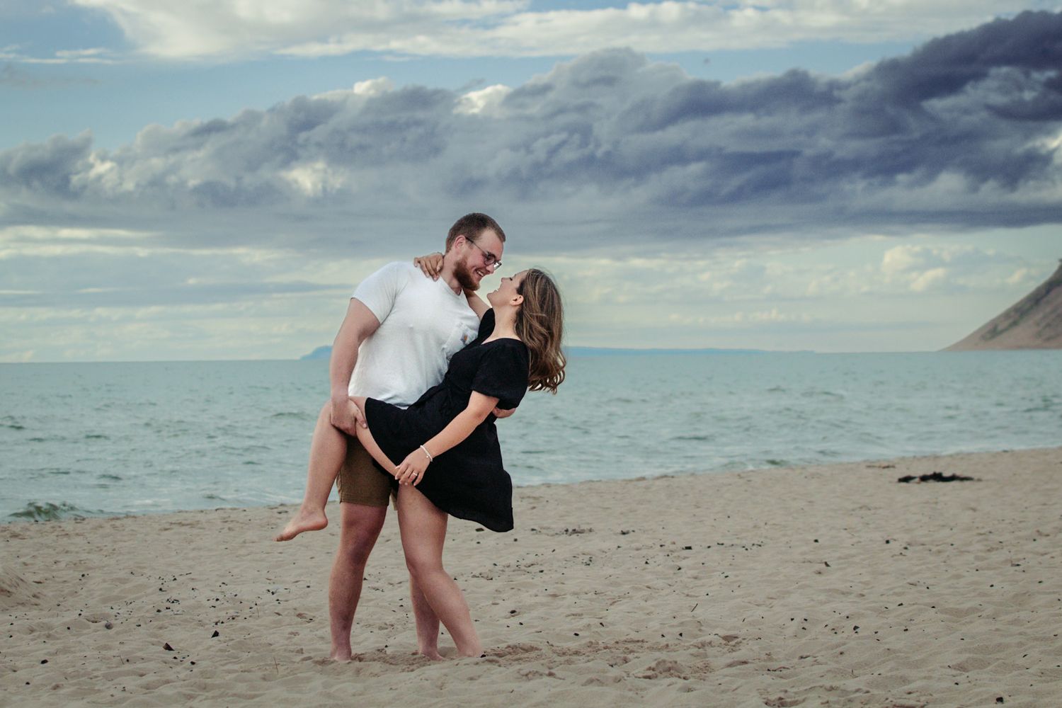 A romantic couple embraces on a scenic beach with cloudy skies and mountains in the background.