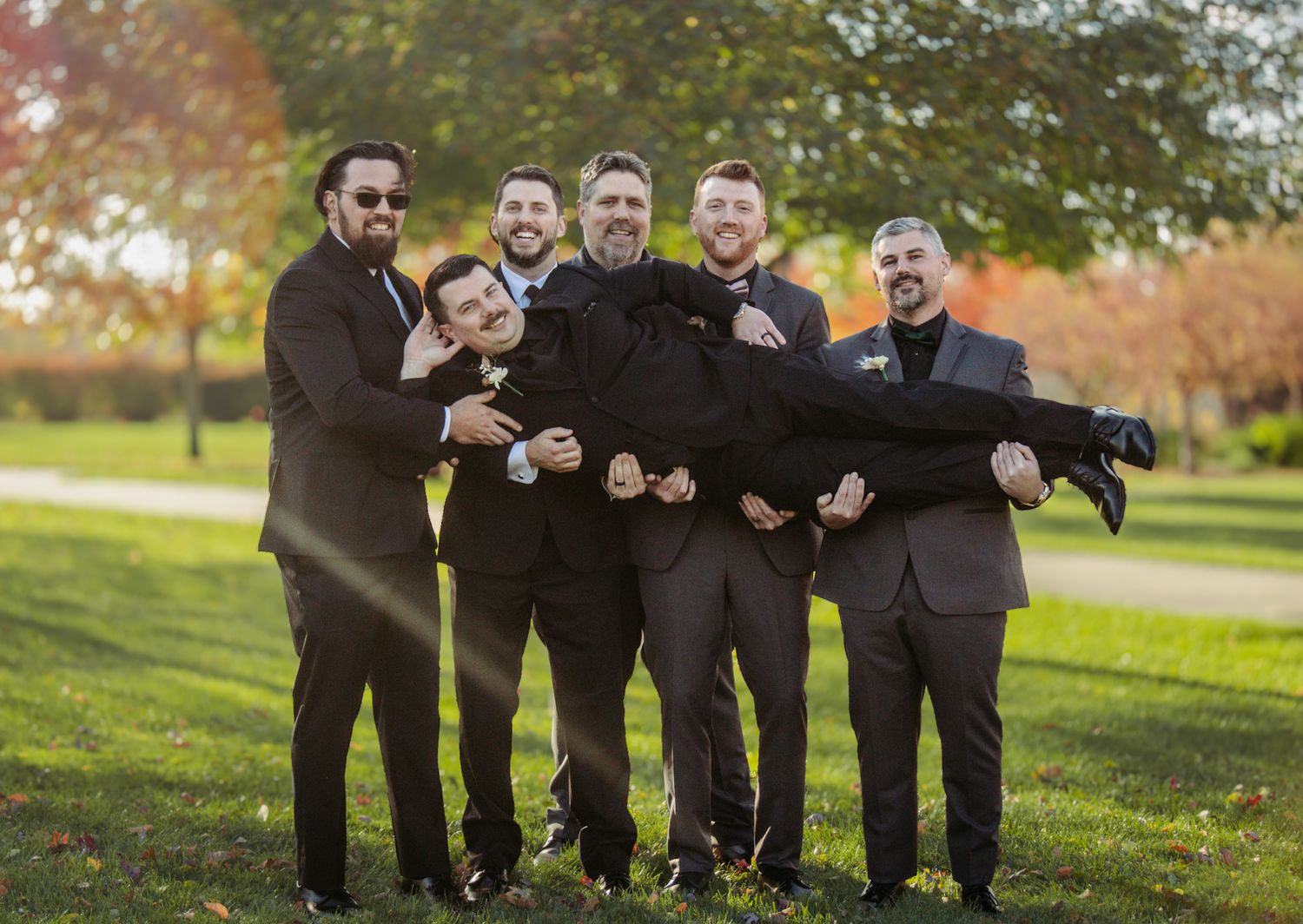 Groomsmen in black suits posing playfully on grass during fall wedding photo session.