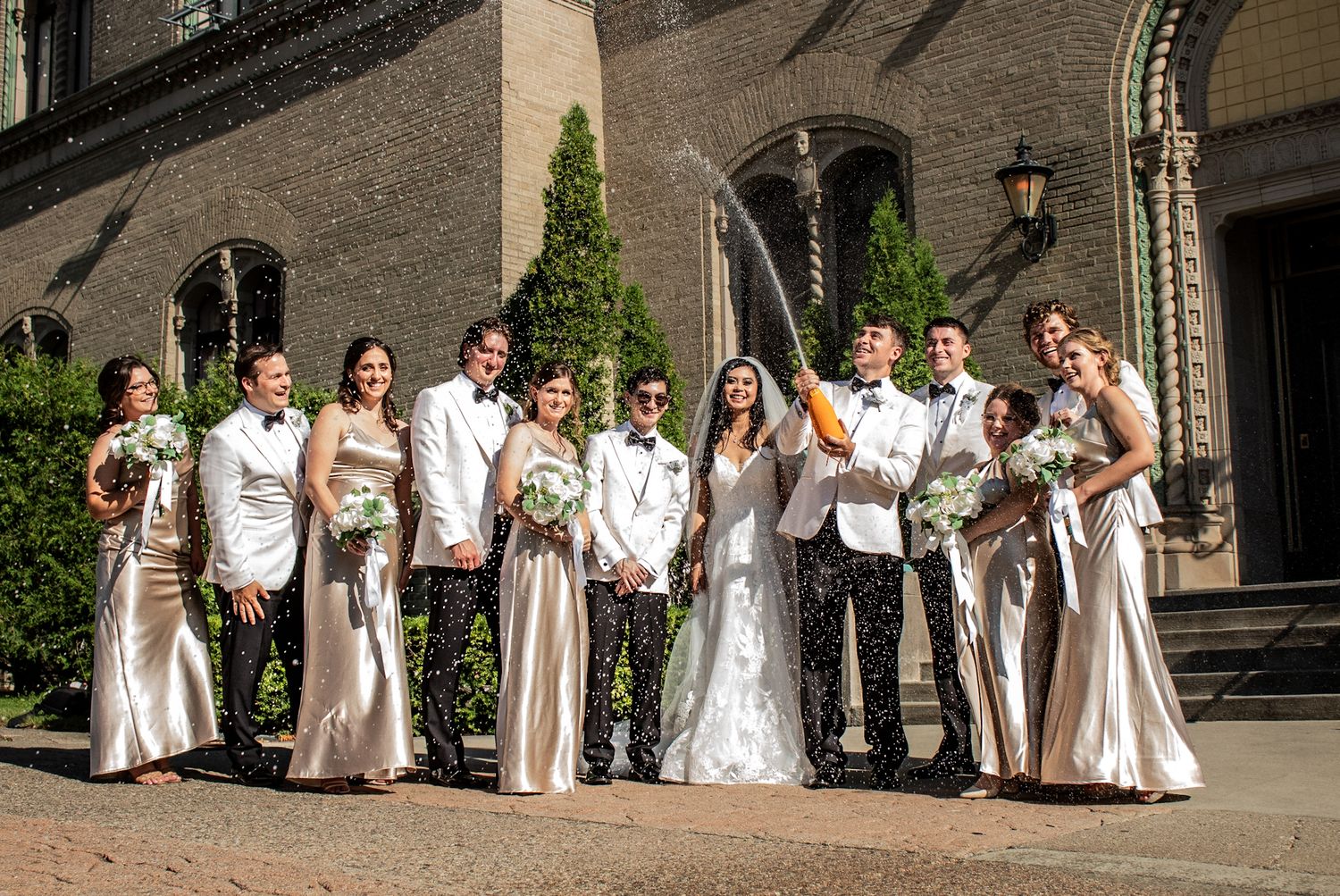 Wedding party in white and cream formal attire poses together outside historic building.