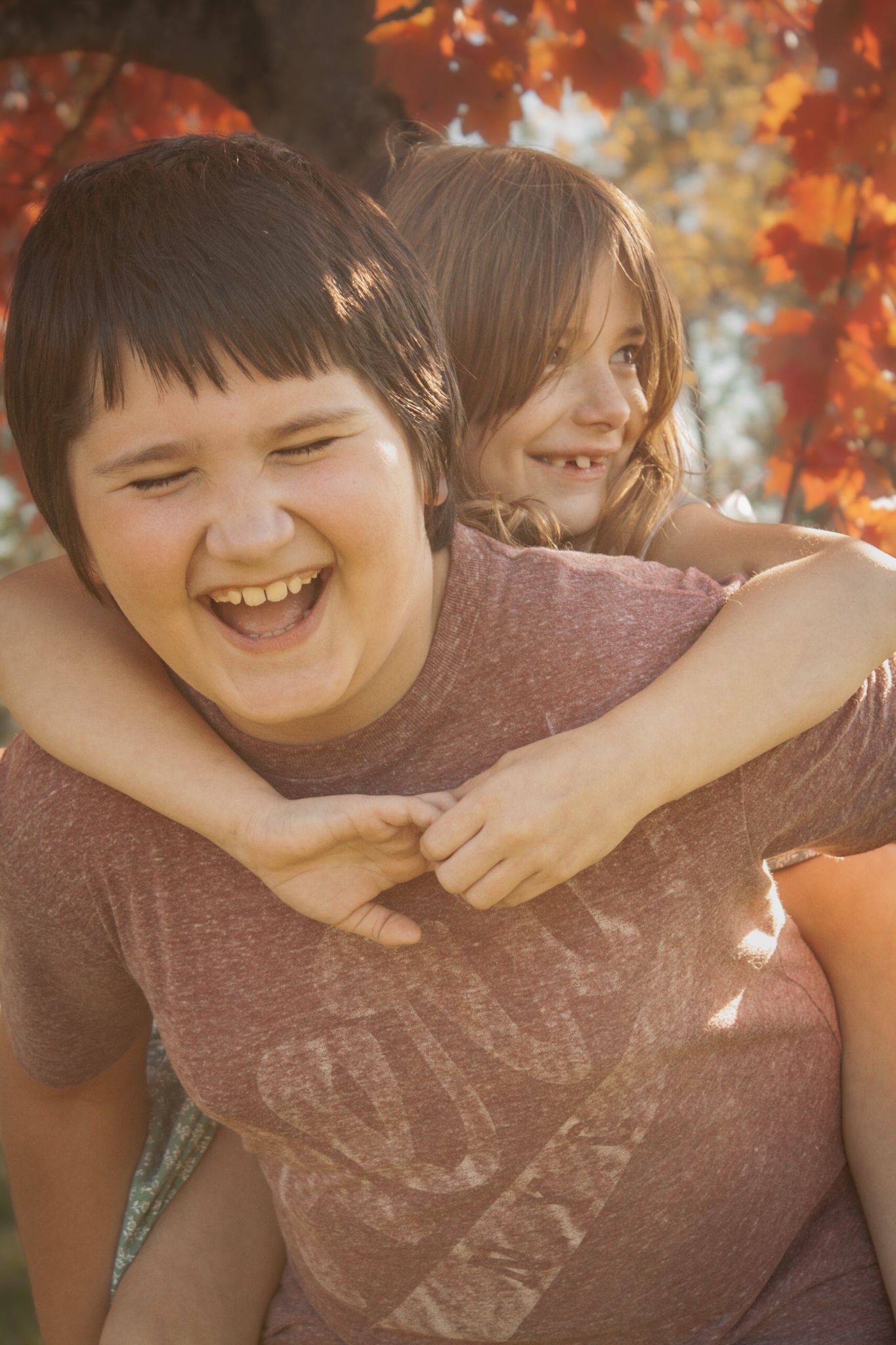 Two children share a playful piggyback ride moment among autumn foliage.