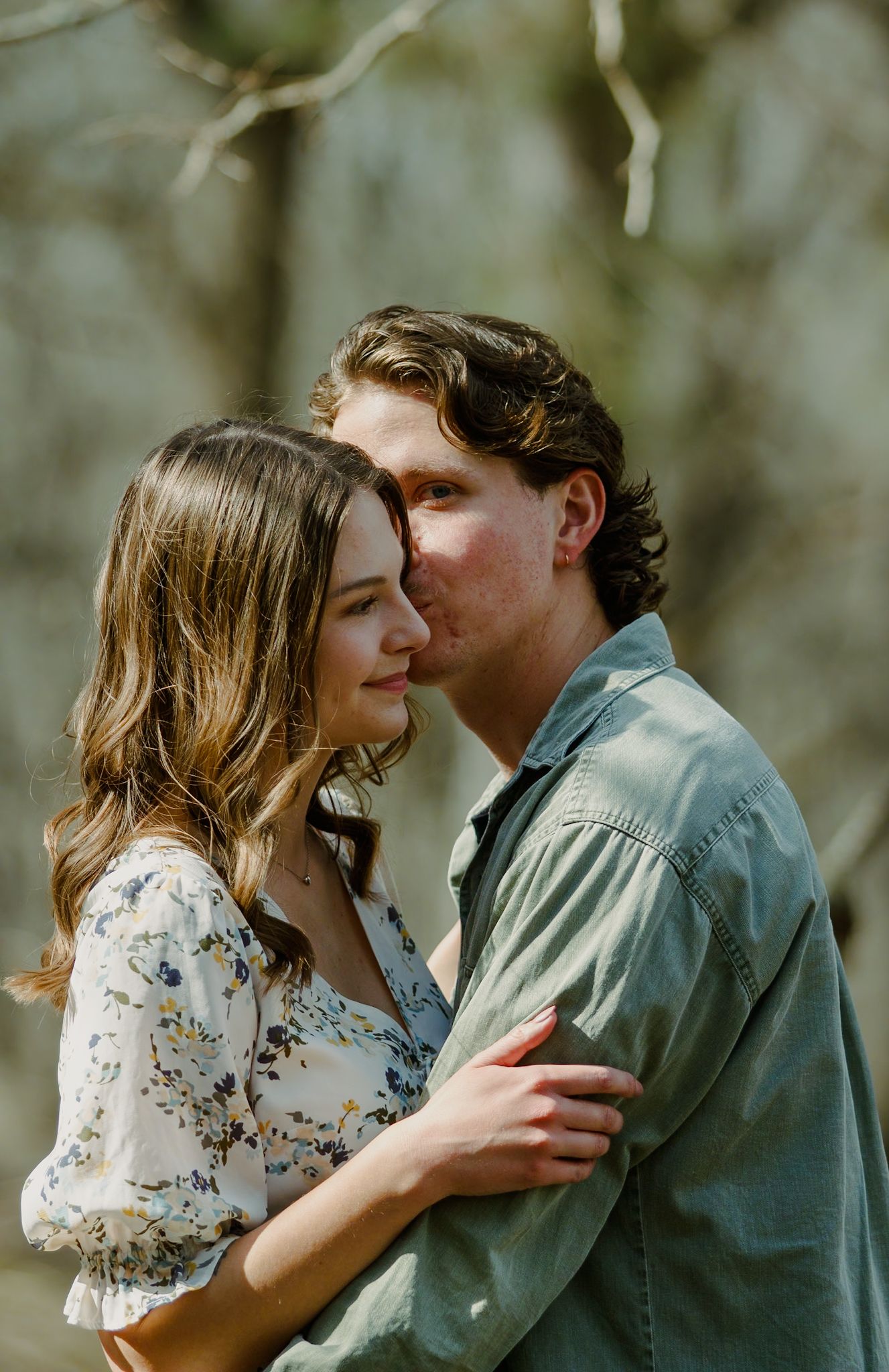 A romantic outdoor portrait shows a couple in tender embrace against a soft natural background.