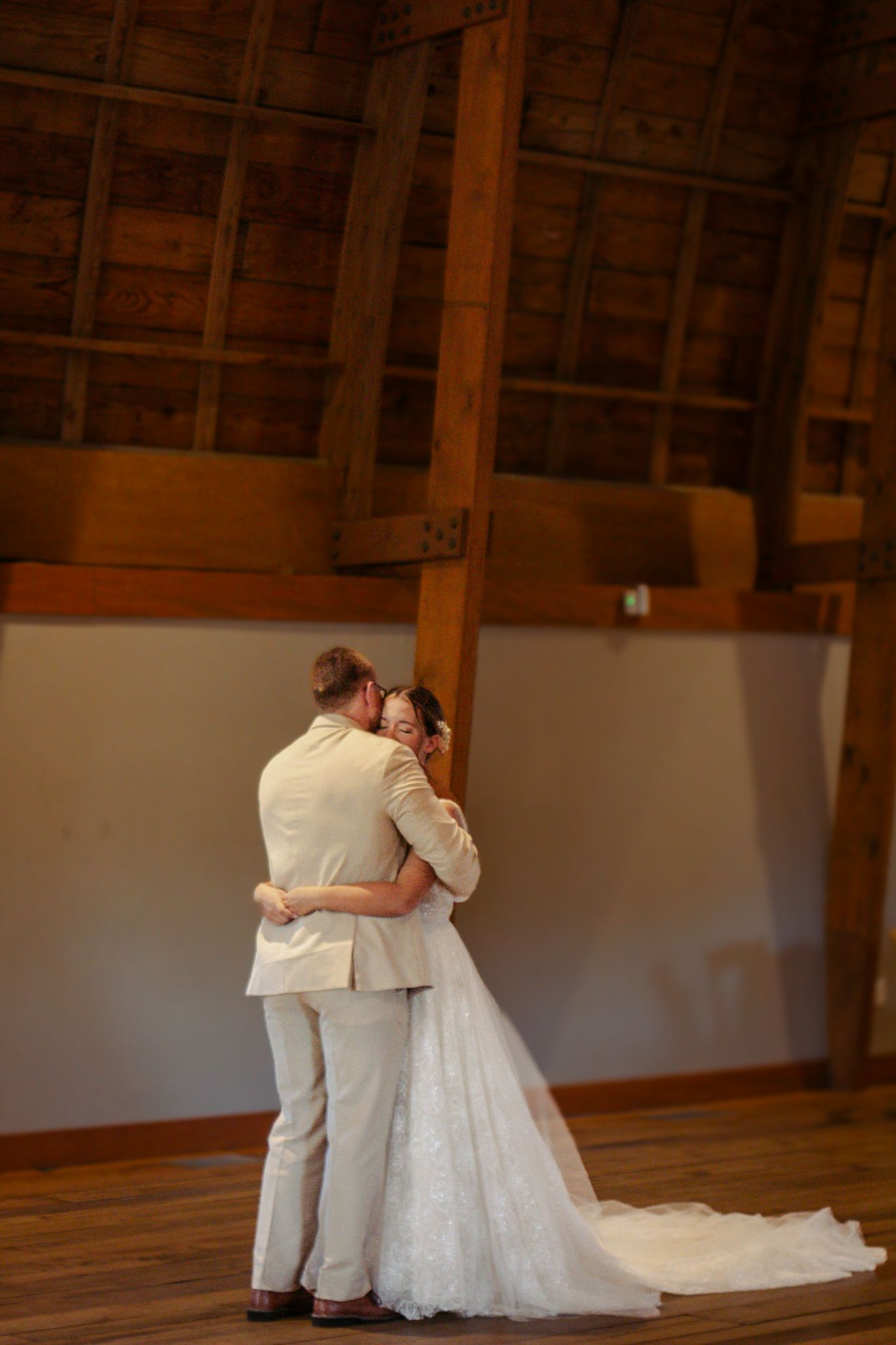 Newlyweds embrace during their first dance on a wooden floor beneath exposed wooden beams at The Cathedral Barn in Traverse City, Michigan.