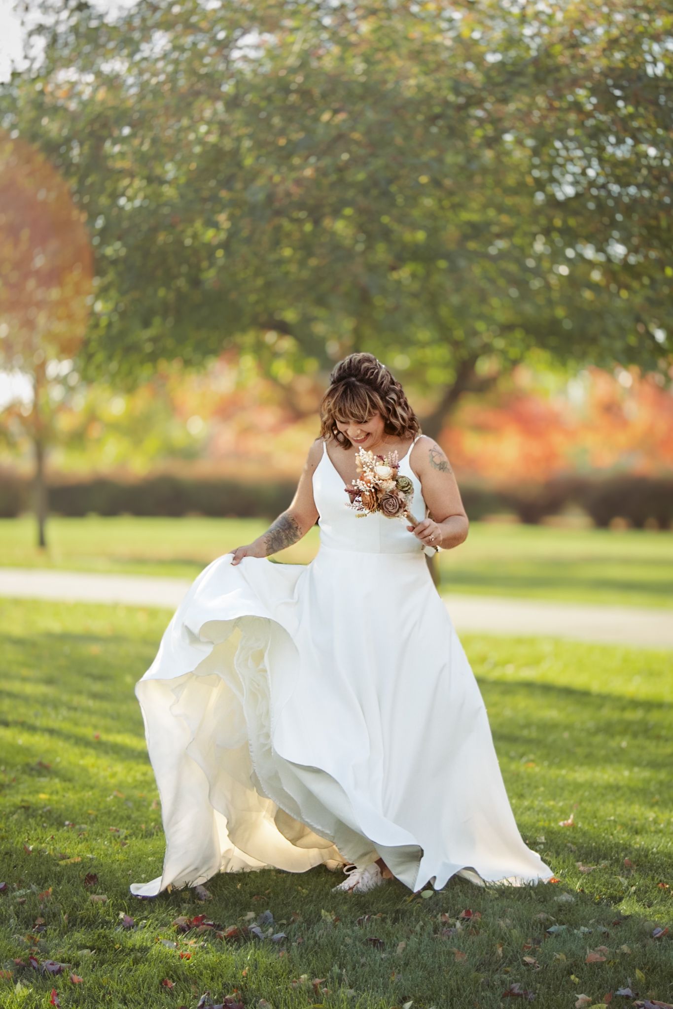 A bride in a flowing white gown twirls on grass with autumn trees in the background.