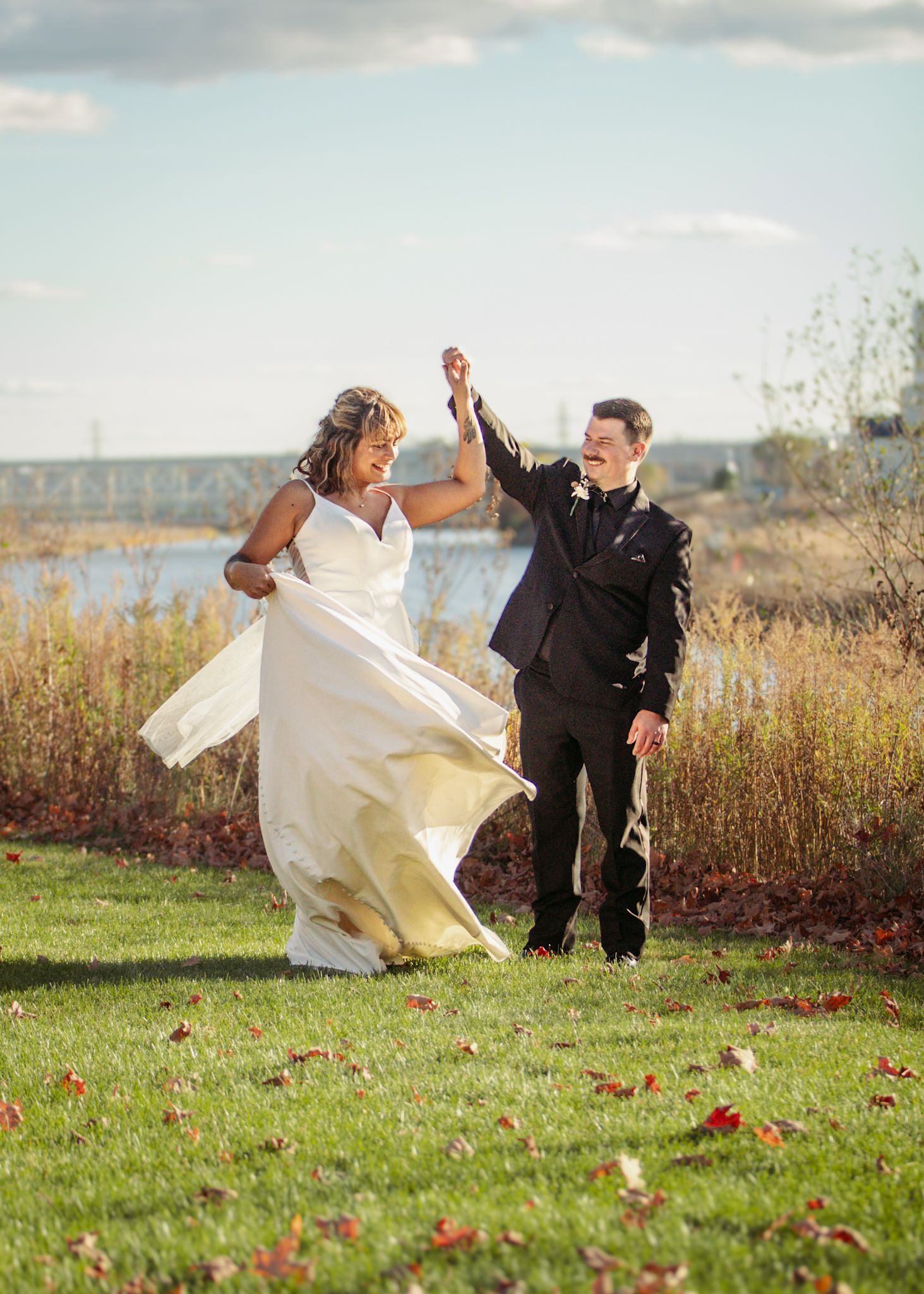 Wedding couple playfully runs through a grassy field with flowing white dress by the water.