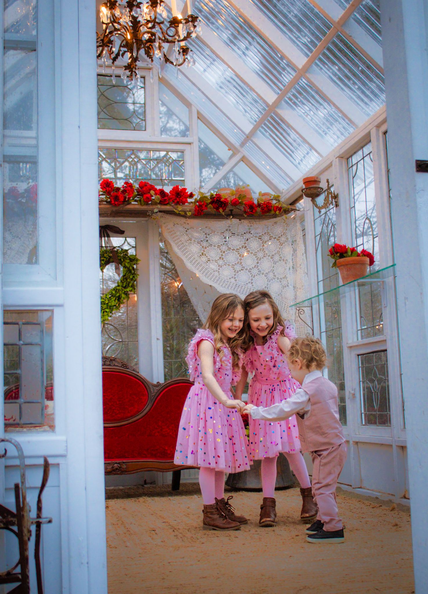 Children in pink outfits playing together inside a sunlit conservatory decorated with red flowers and chandeliers.