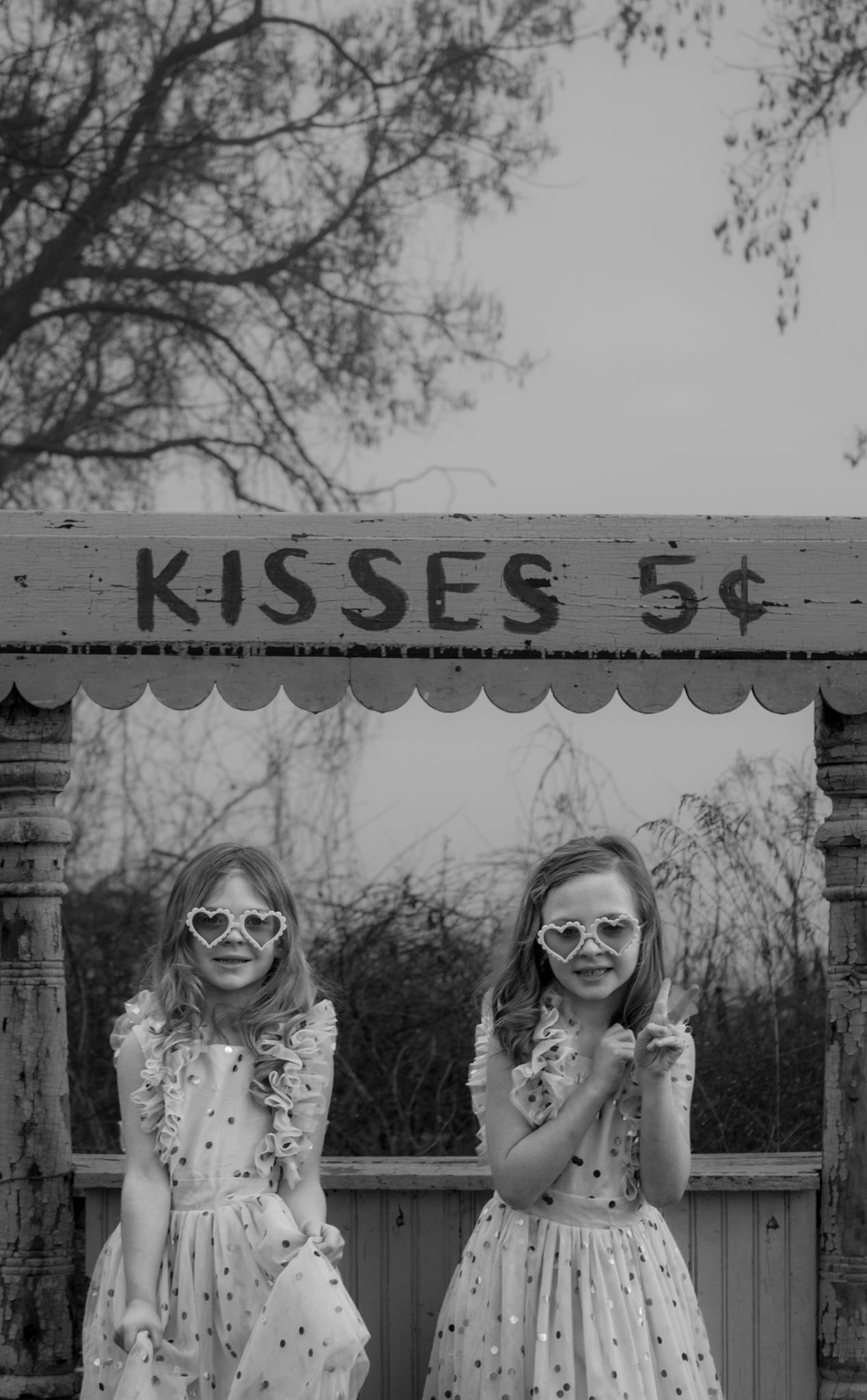 Vintage style photo of two children in sunglasses posing under 'Kisses 5 cents' sign.