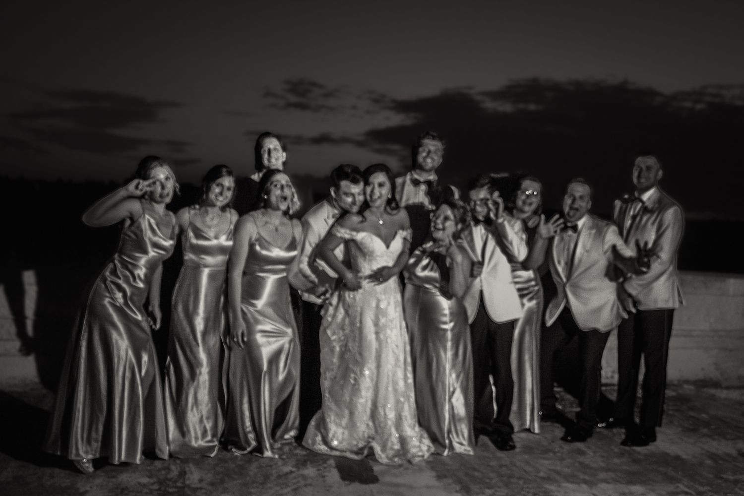 Black and white panoramic photo of a wedding party posing together outdoors at dusk.