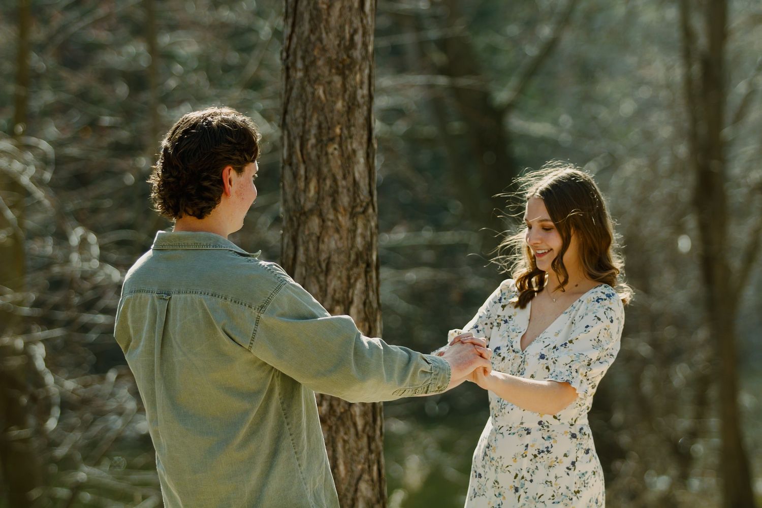 A couple holds hands while dancing in a sunlit forest setting.