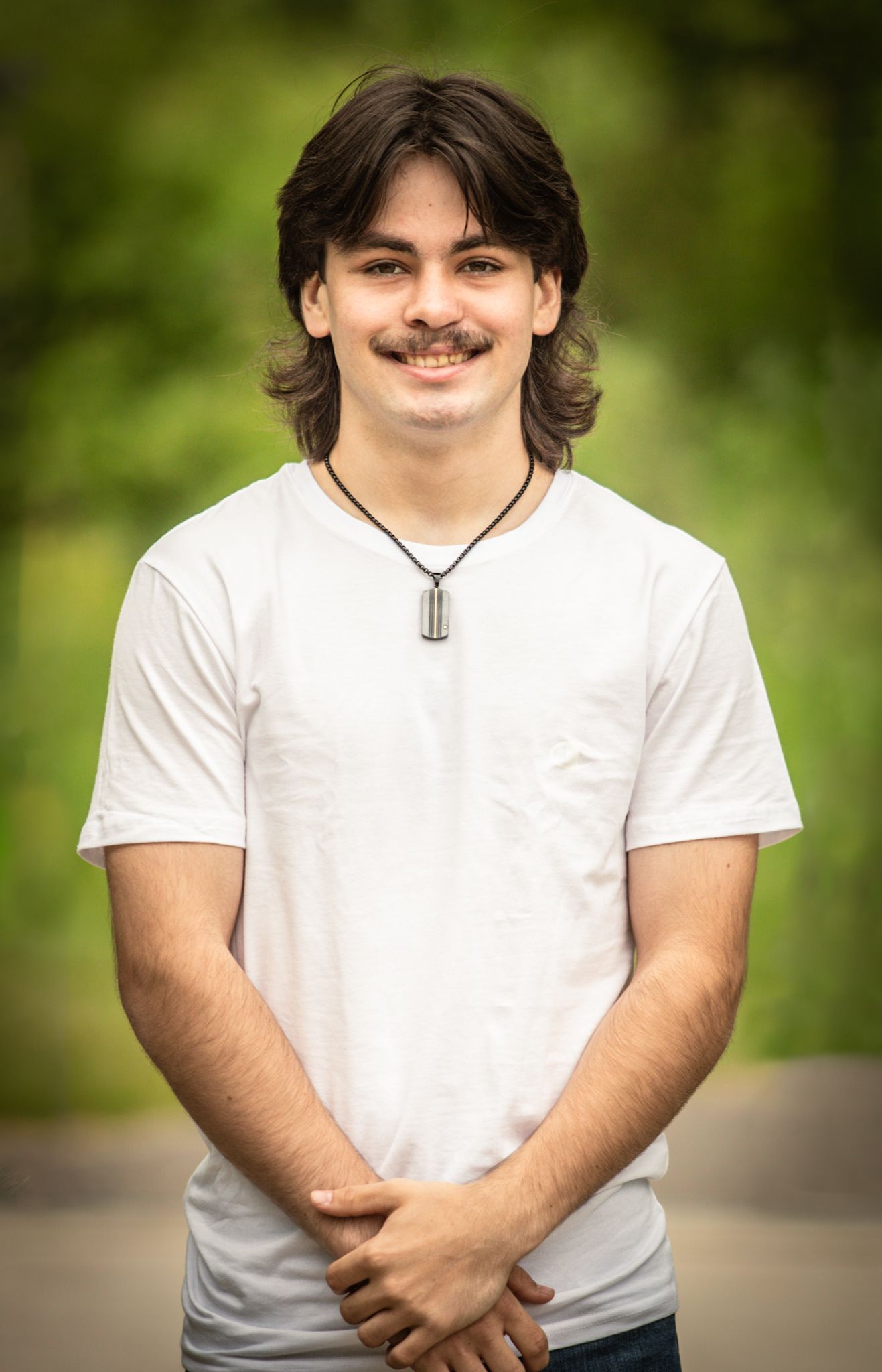 A person in a white t-shirt and necklace poses against a blurred green nature background.