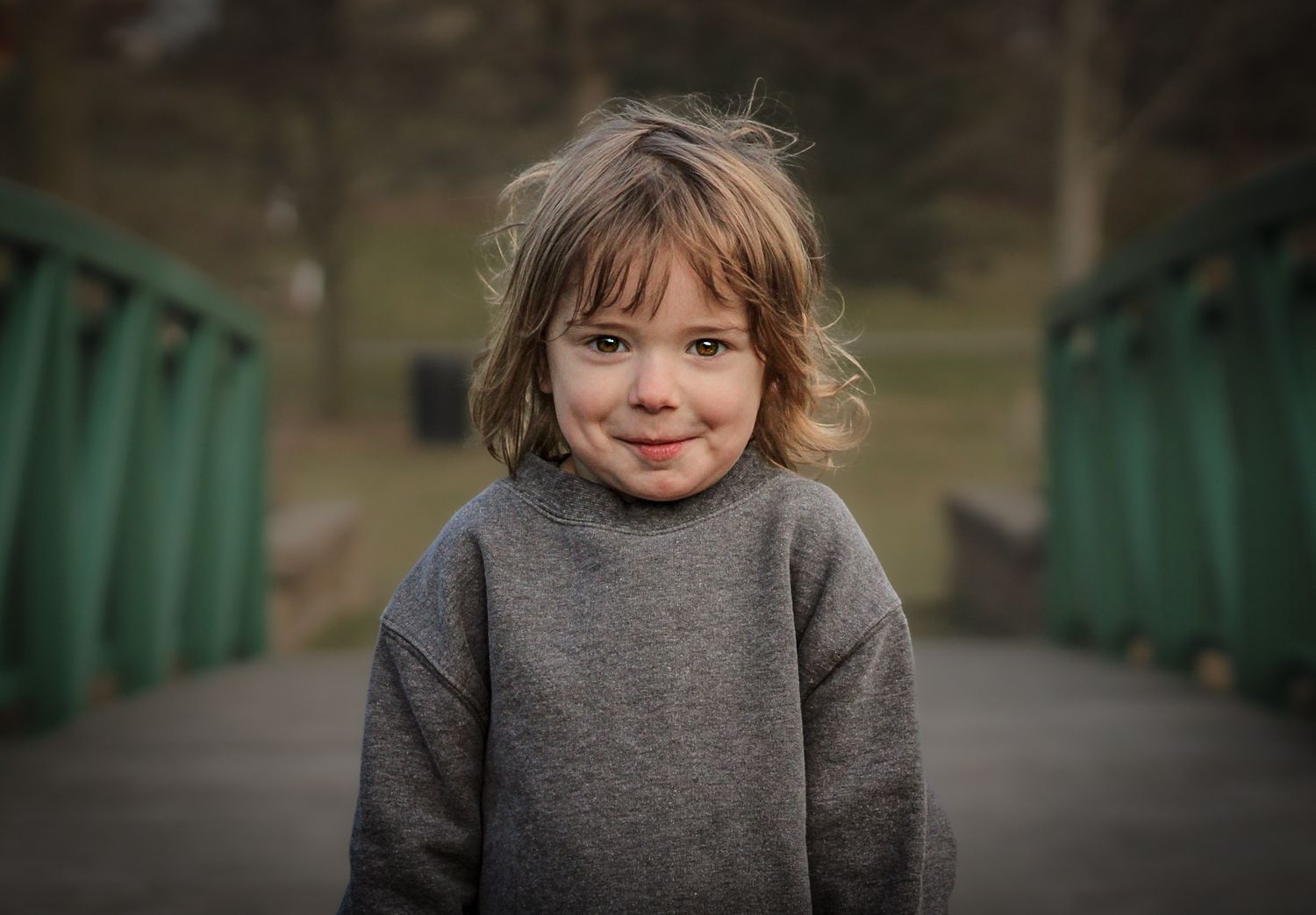 Child in grey sweater smiling on green bridge during outdoor portrait session.