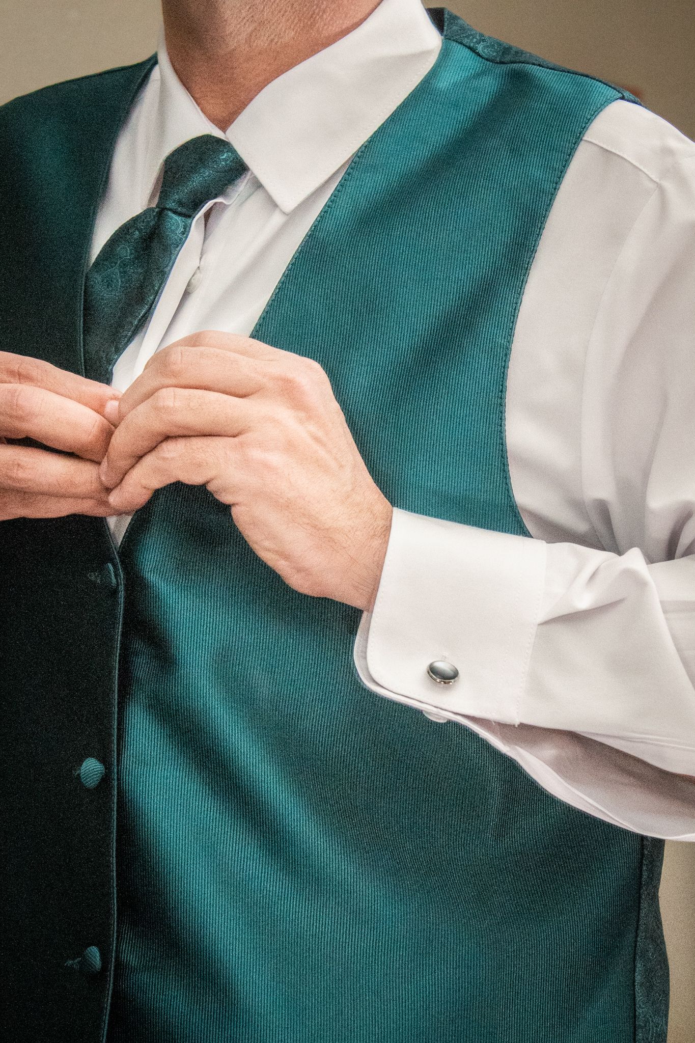 Person adjusting a teal silk vest over white dress shirt with french cuffs.