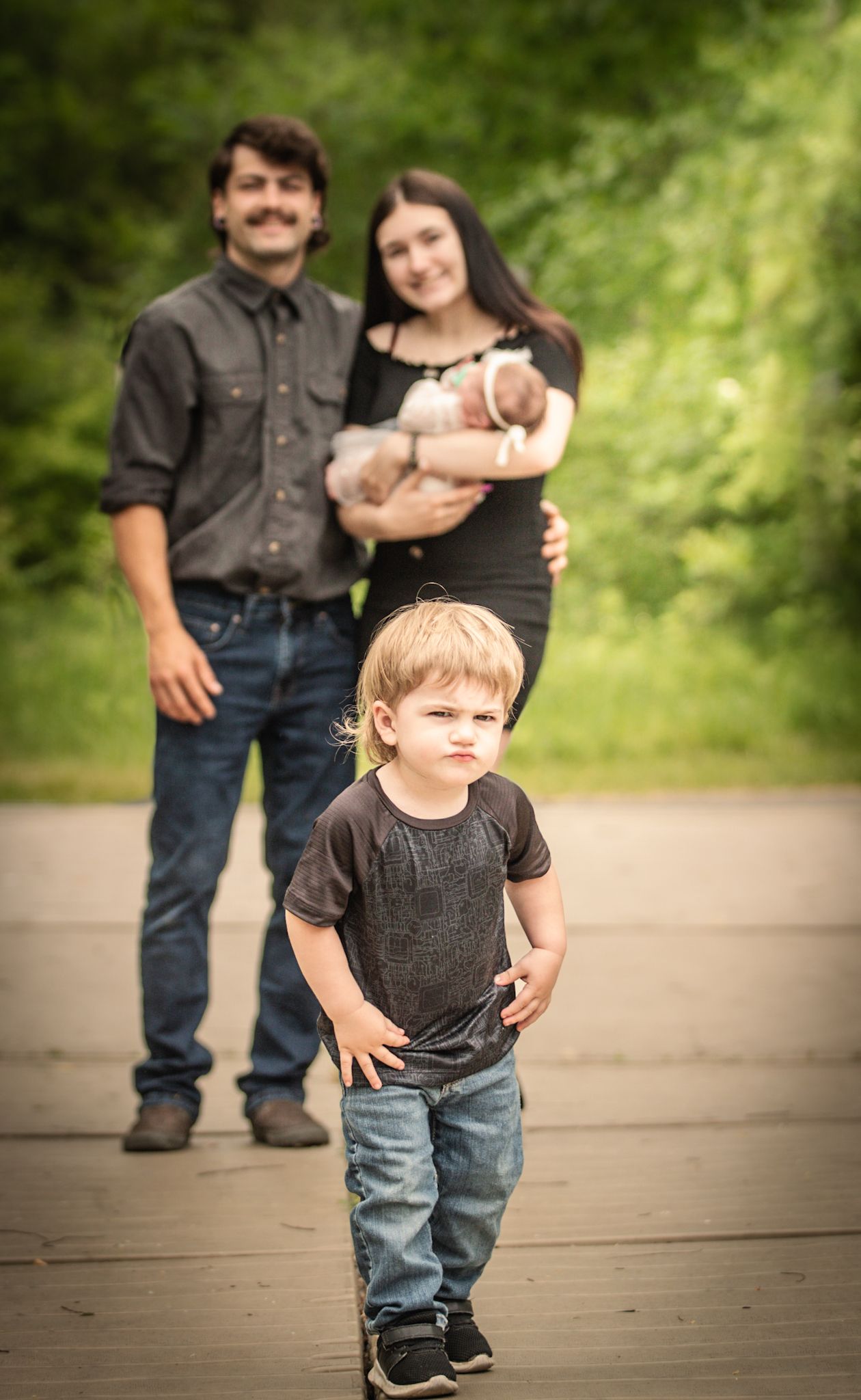 A toddler in jeans stands in foreground while family members hold a baby behind on a paved path.