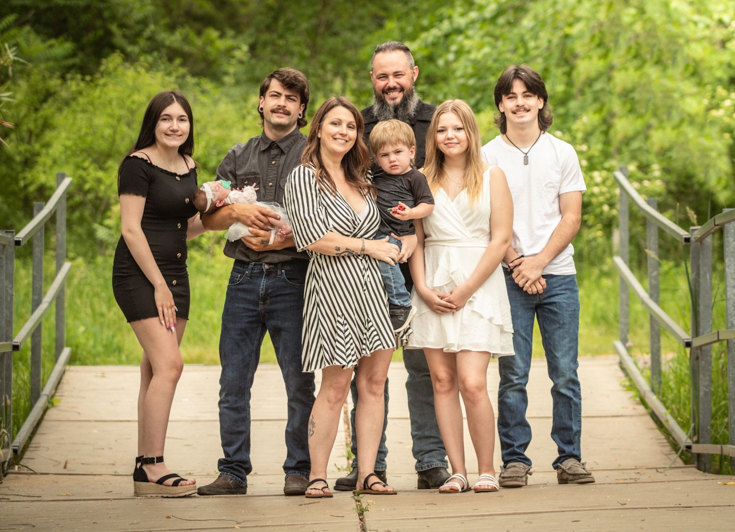 Happy family of seven poses together on a wooden bridge surrounded by lush greenery.