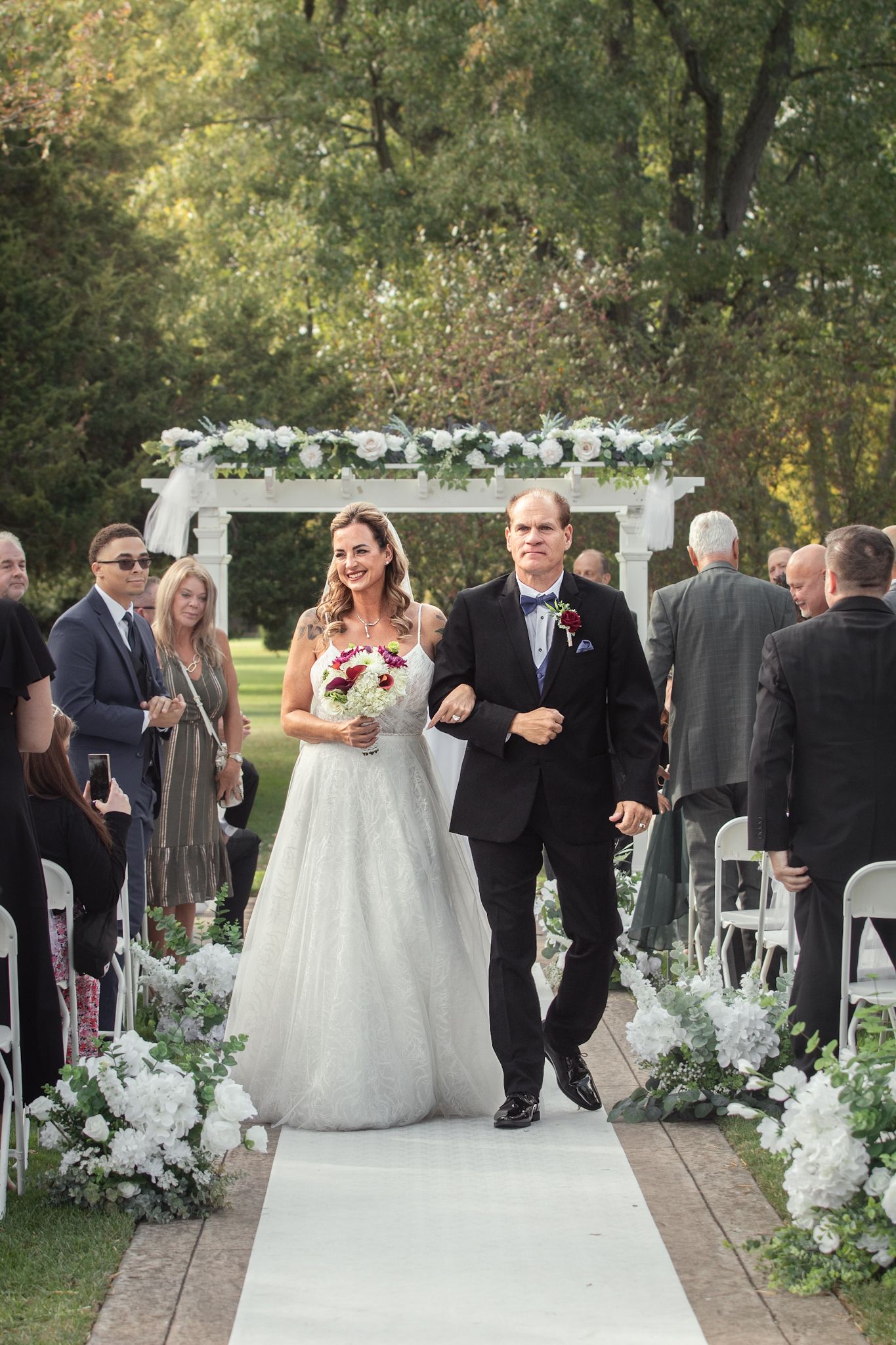 Elegant outdoor wedding ceremony setup with white floral arrangements and guests gathering.