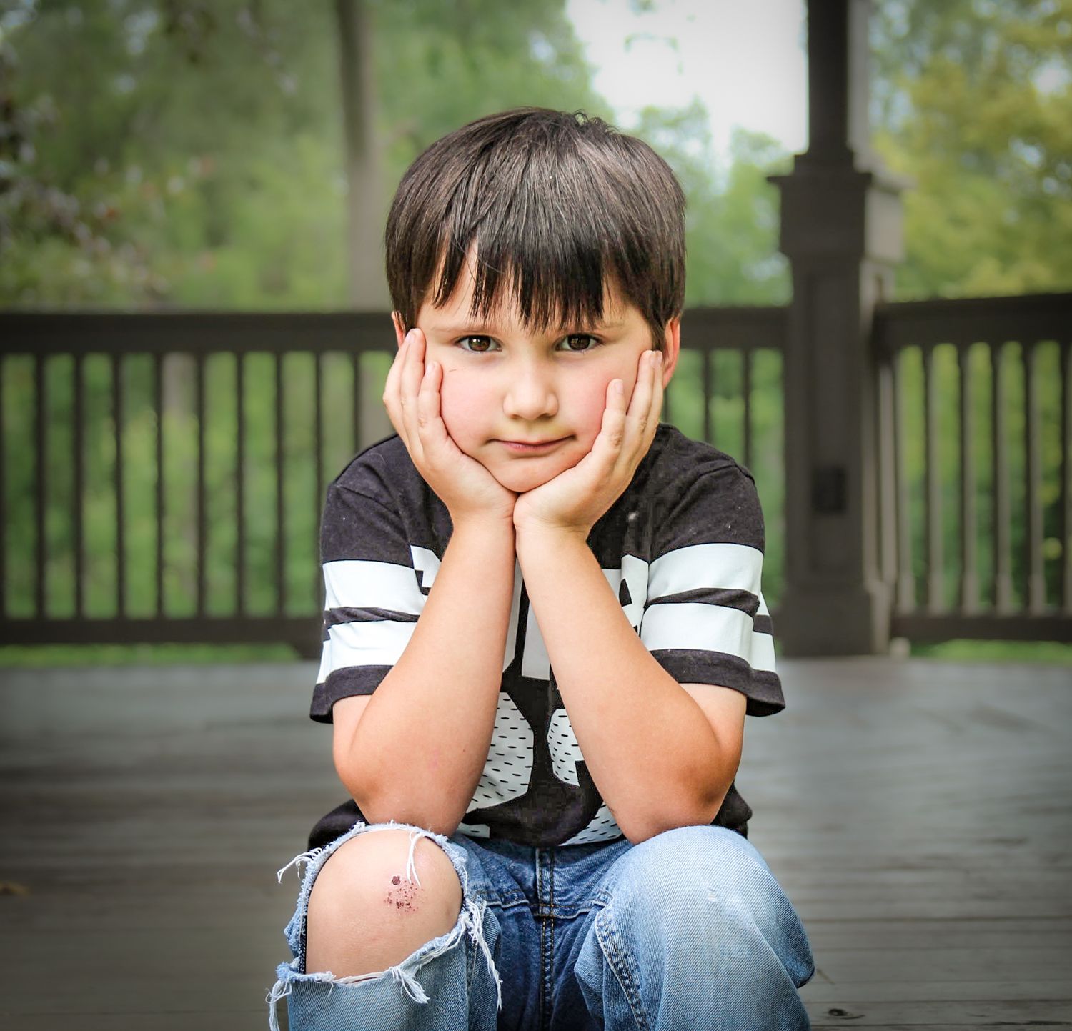 Portrait of someone sitting on porch with hands on cheeks wearing striped shirt and ripped jeans.