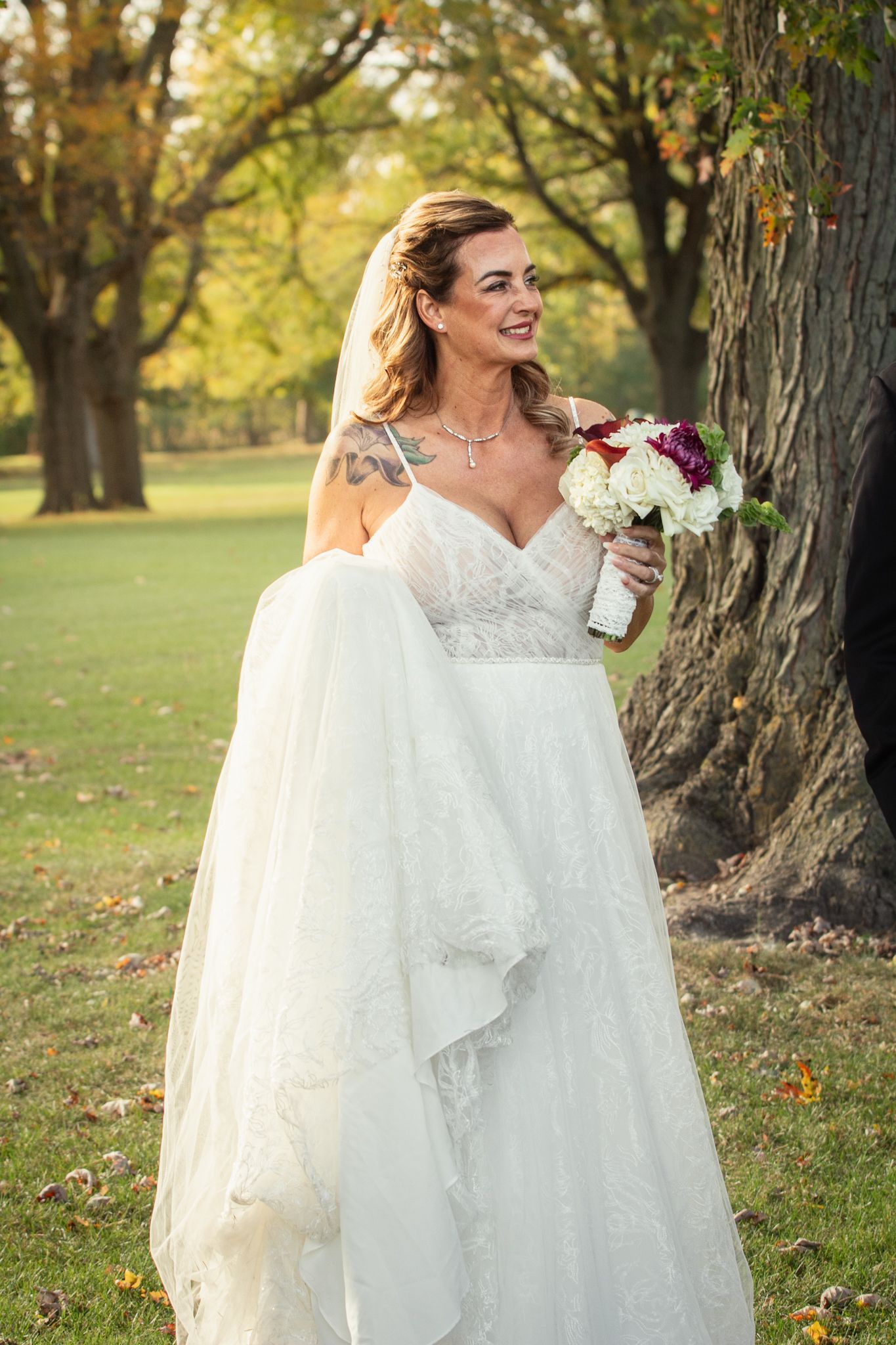 A bride in a white lace gown laughs while holding a bouquet in an autumn park setting.