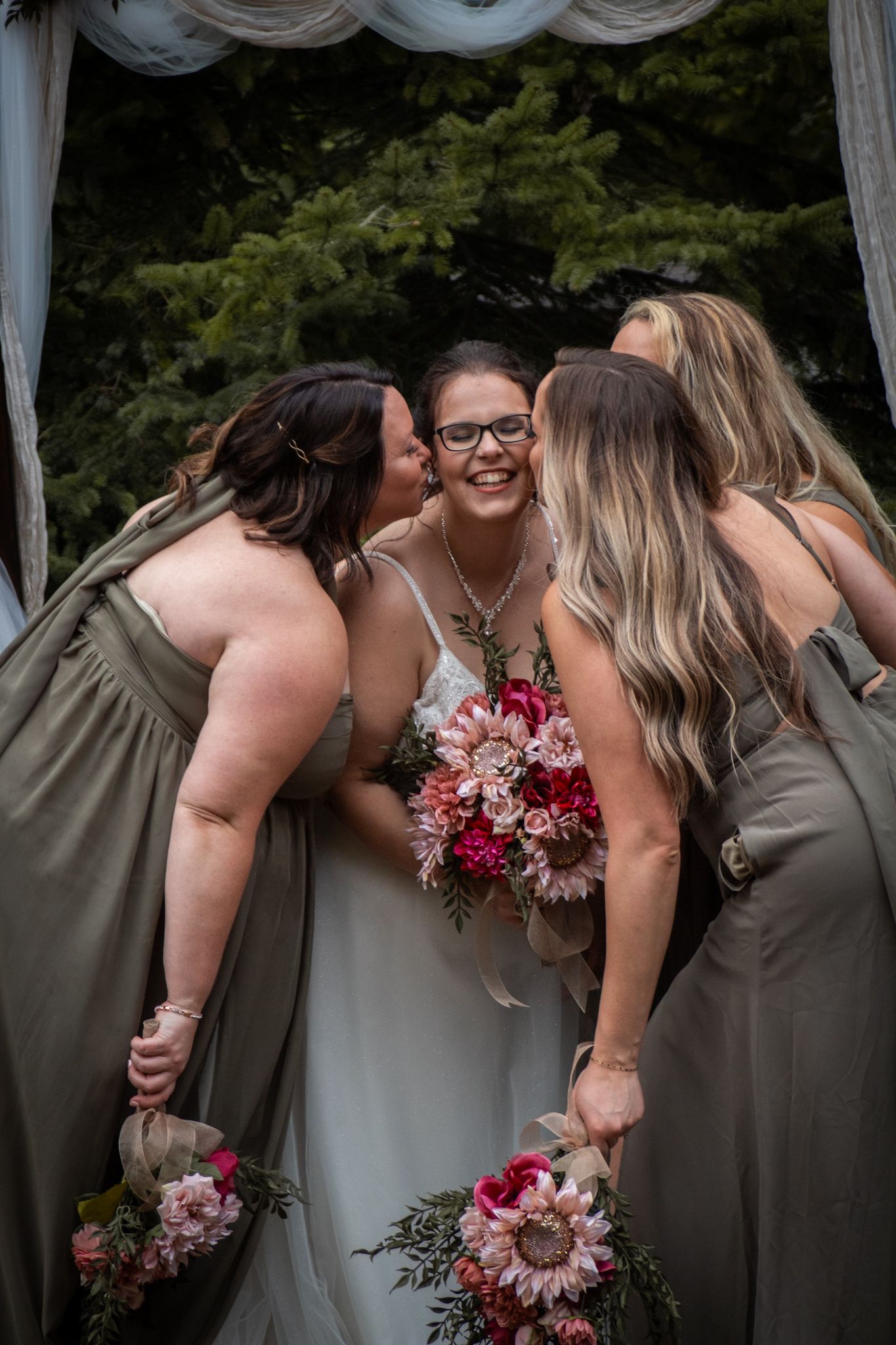 Wedding party shares an intimate moment under a decorated arch with evergreen trees in background.