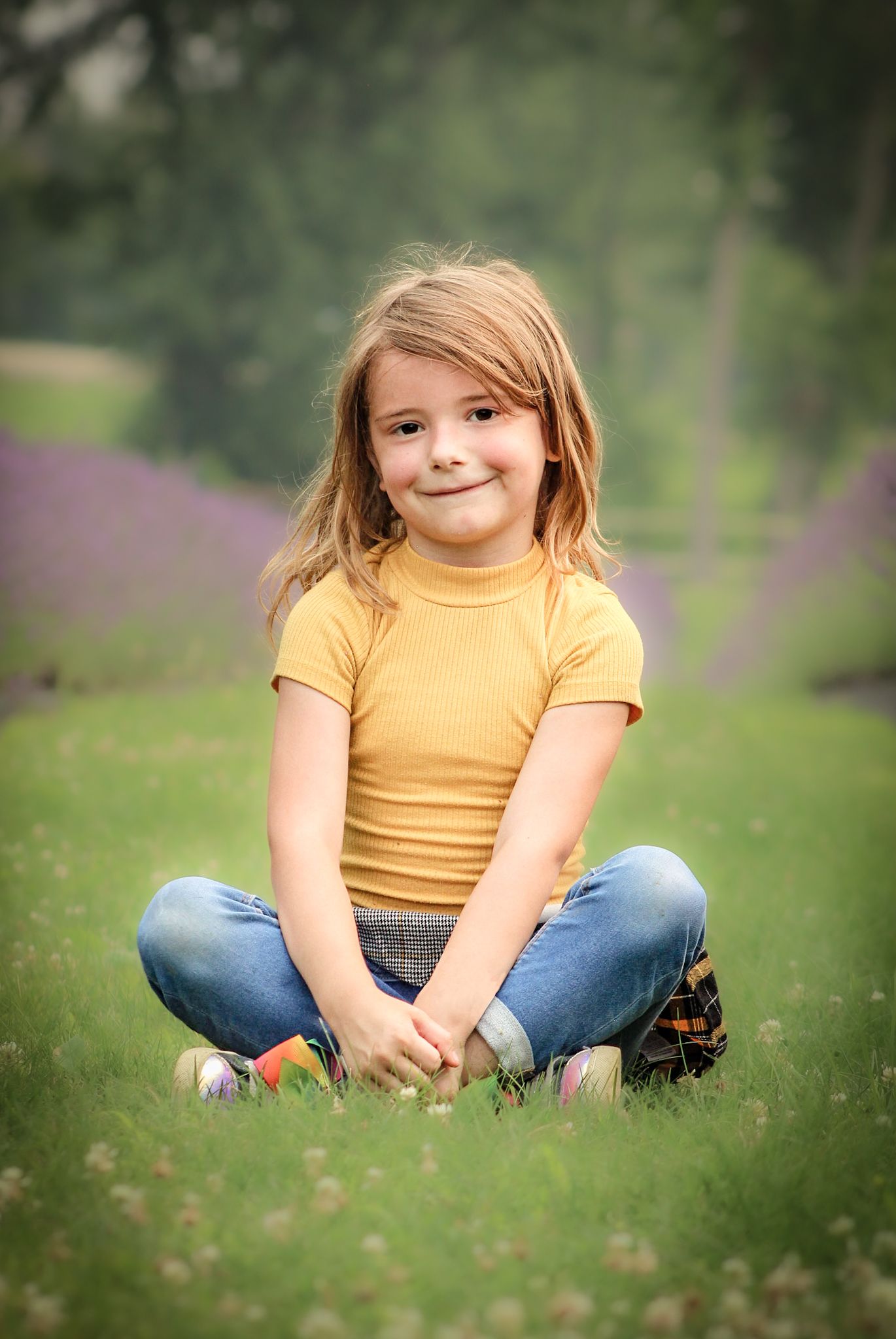 Child in yellow shirt and jeans sits cross-legged on grass in garden setting.