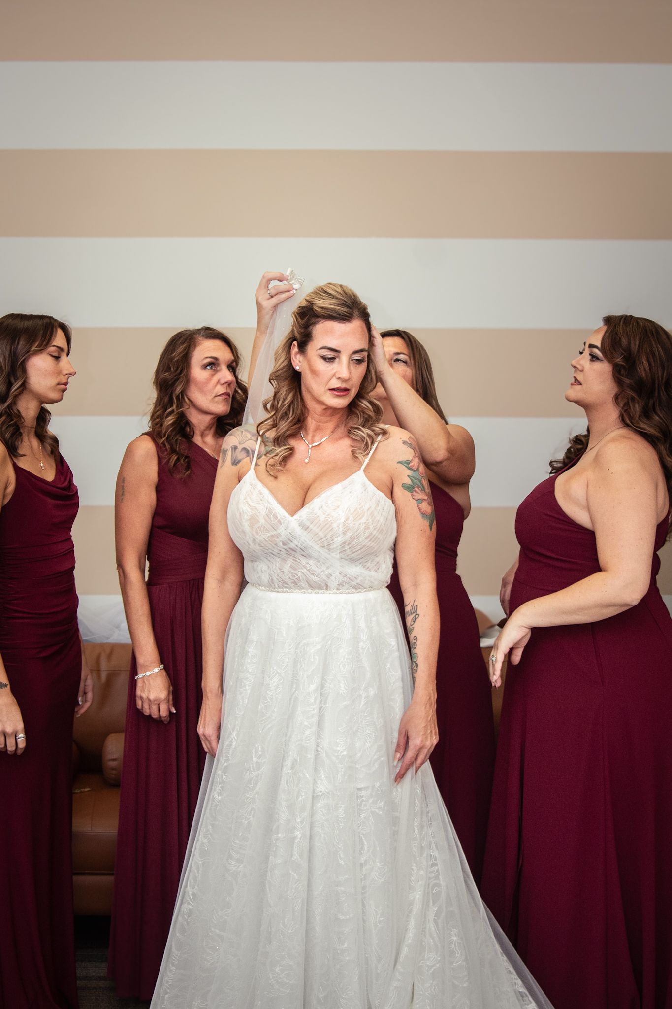 Bride in white lace dress poses with bridesmaids wearing burgundy dresses during wedding preparations.