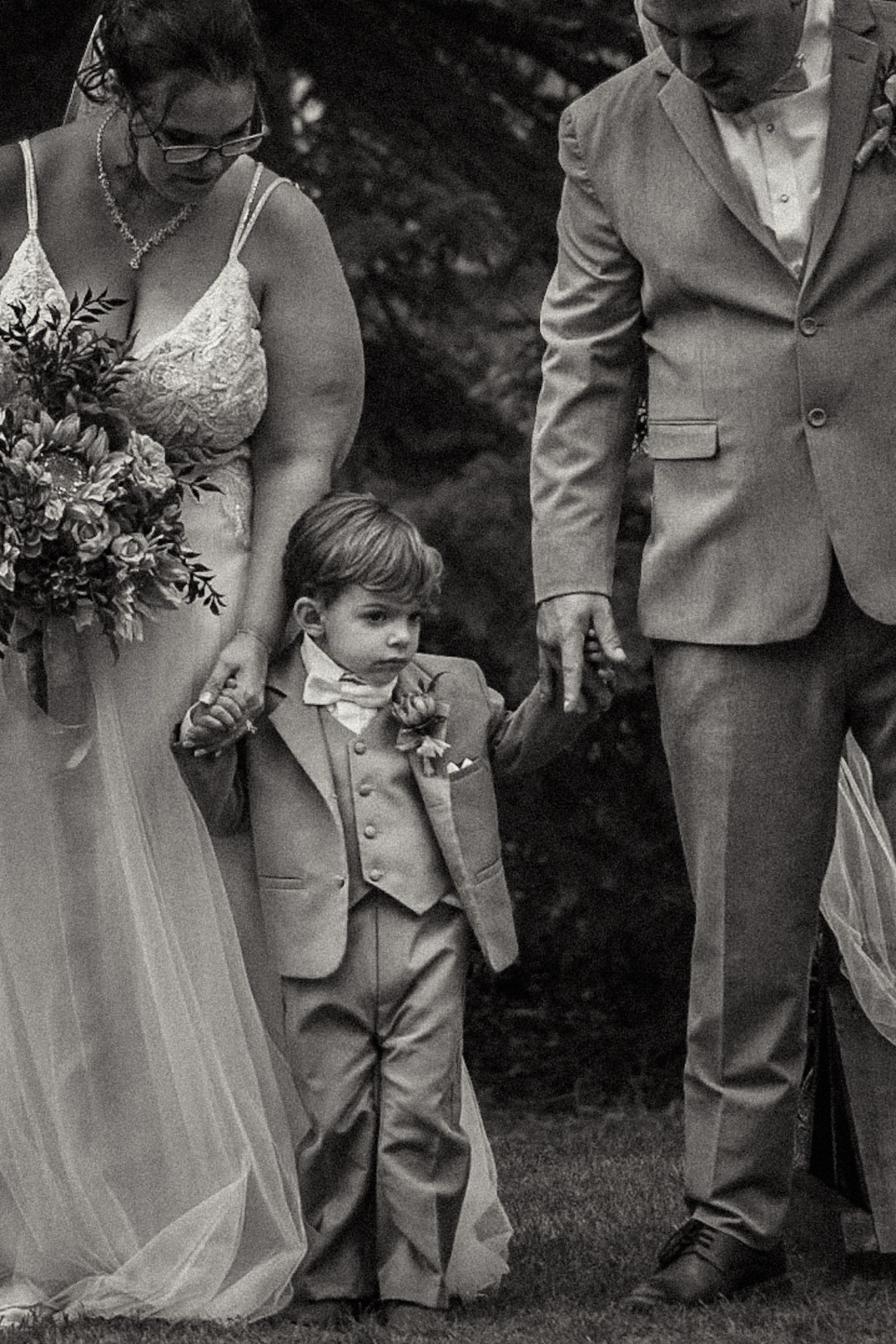 A black and white photo of a small child in formal wear walking with adults at a wedding.