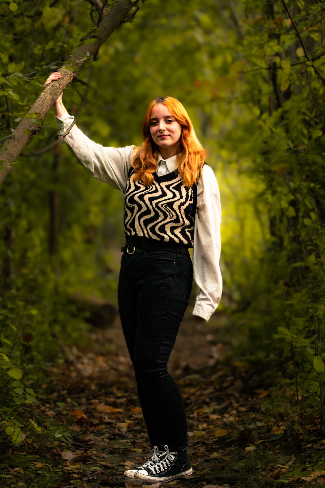 A person in a zigzag pattern sweater and black pants poses playfully on a wooded trail.