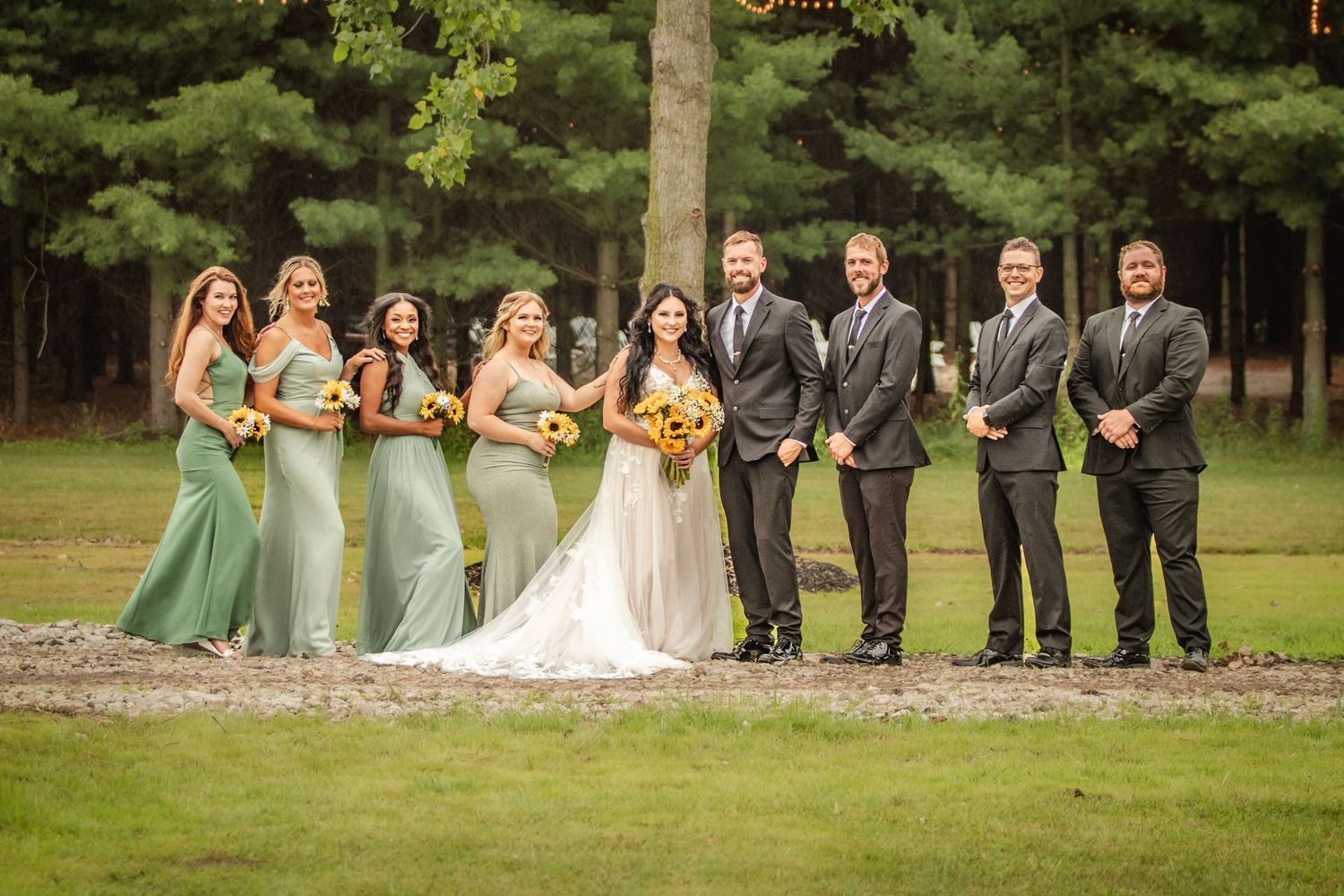 Wedding party poses in sage green and black attire on lush garden grounds.