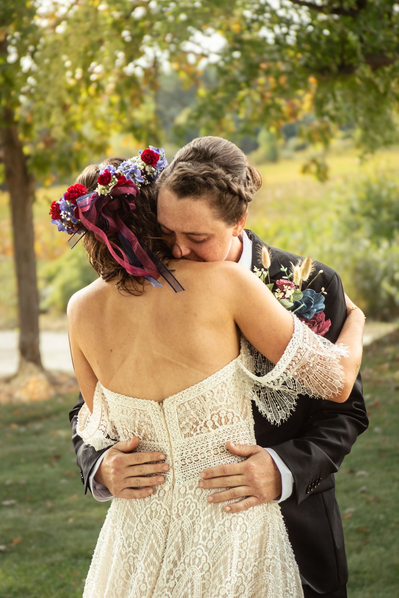 Bride in lace dress with floral crown embraced from behind during outdoor autumn wedding.